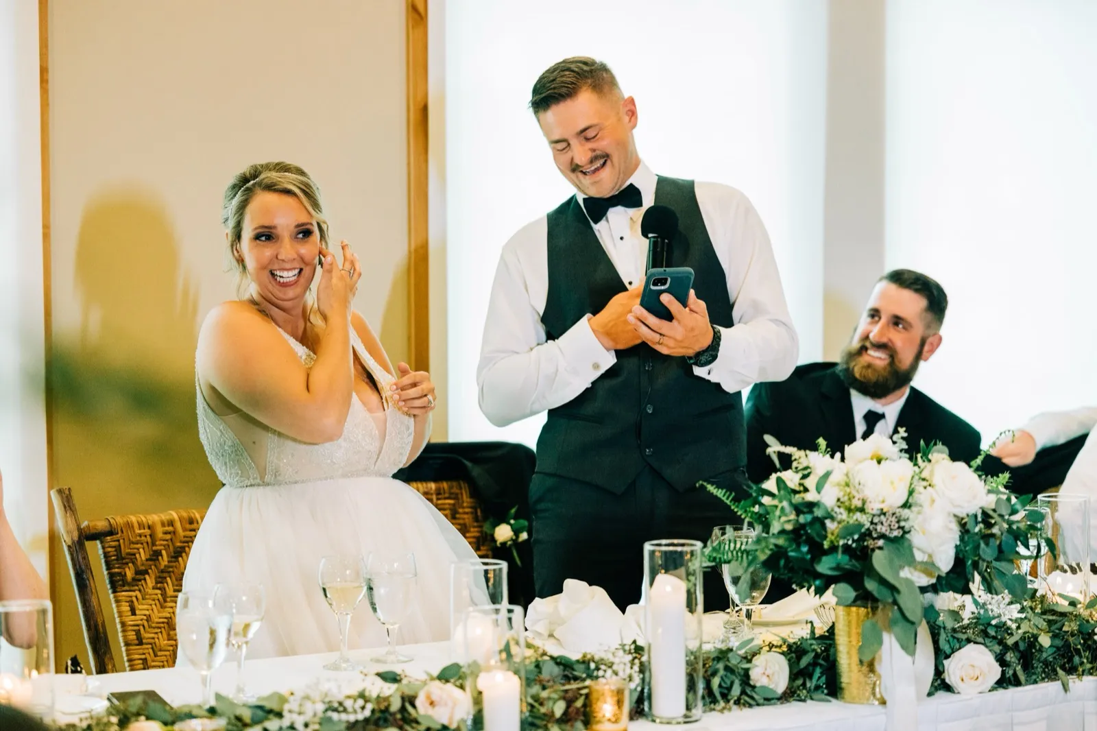 Jonathon grins reading his speech from his phone as Hailee laughs and wipes a tear at the head table — Tim Larsen Photography, Brainerd Lakes MN