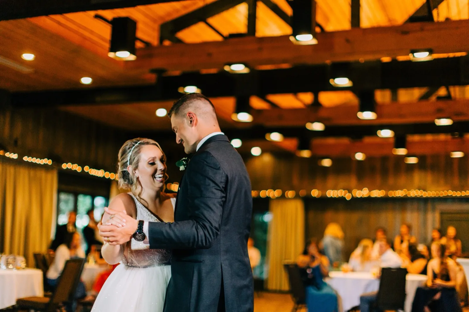 Hailee laughs during the first dance under the exposed beams and cafe lights of the Madden's reception — Tim Larsen Photography, Brainerd Lakes MN