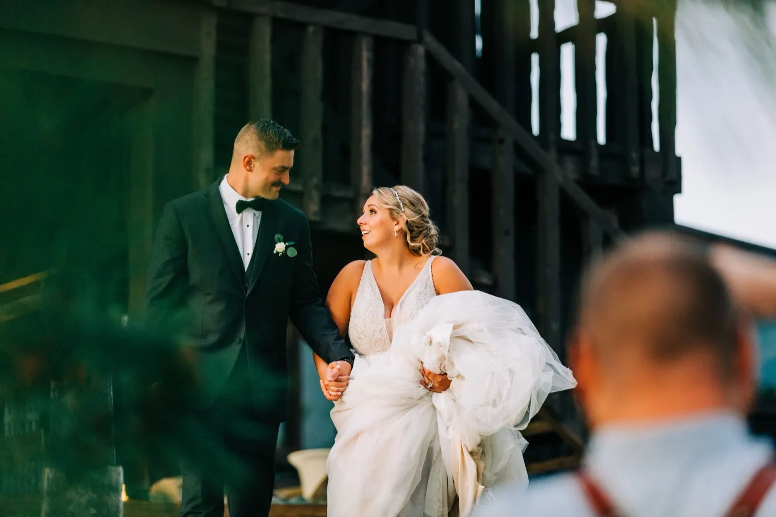 Hailee lifts her gown and looks up at Jonathon, laughing, as they walk hand in hand past a wooden staircase — Tim Larsen Photography, Brainerd Lakes MN