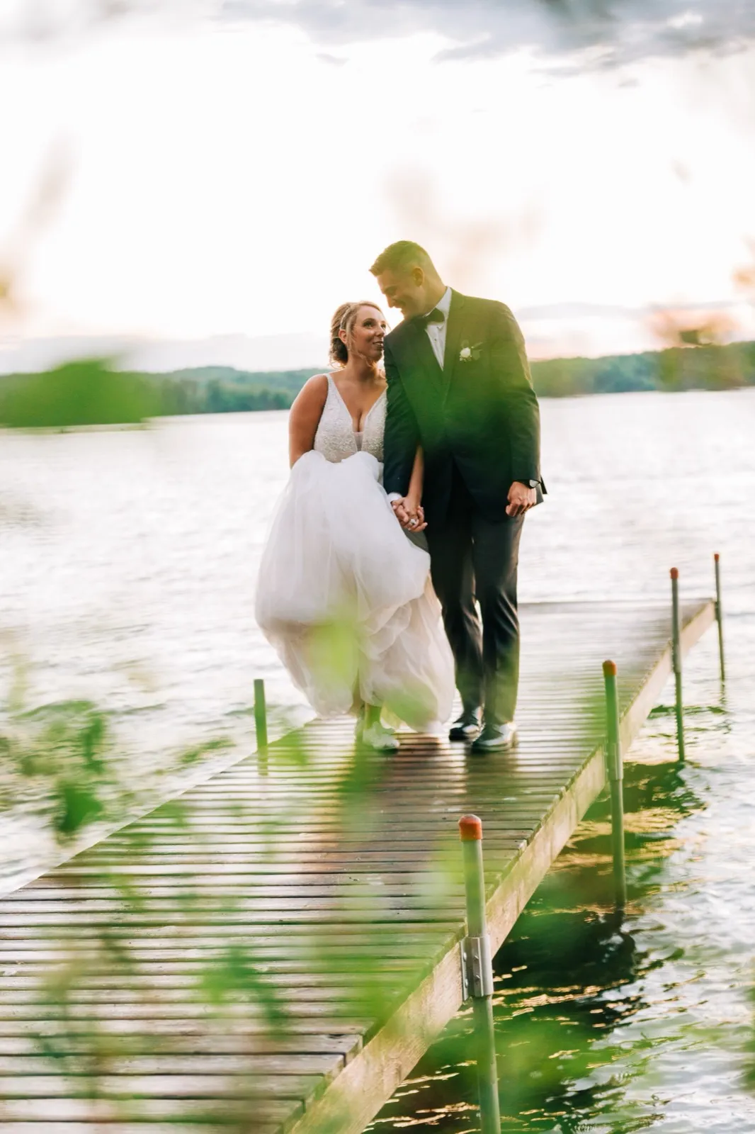 Hailee lifts her tulle skirt as she and Jonathon walk hand in hand down the dock at sunset, the lake backlit behind them — Tim Larsen Photography, Brainerd Lakes MN