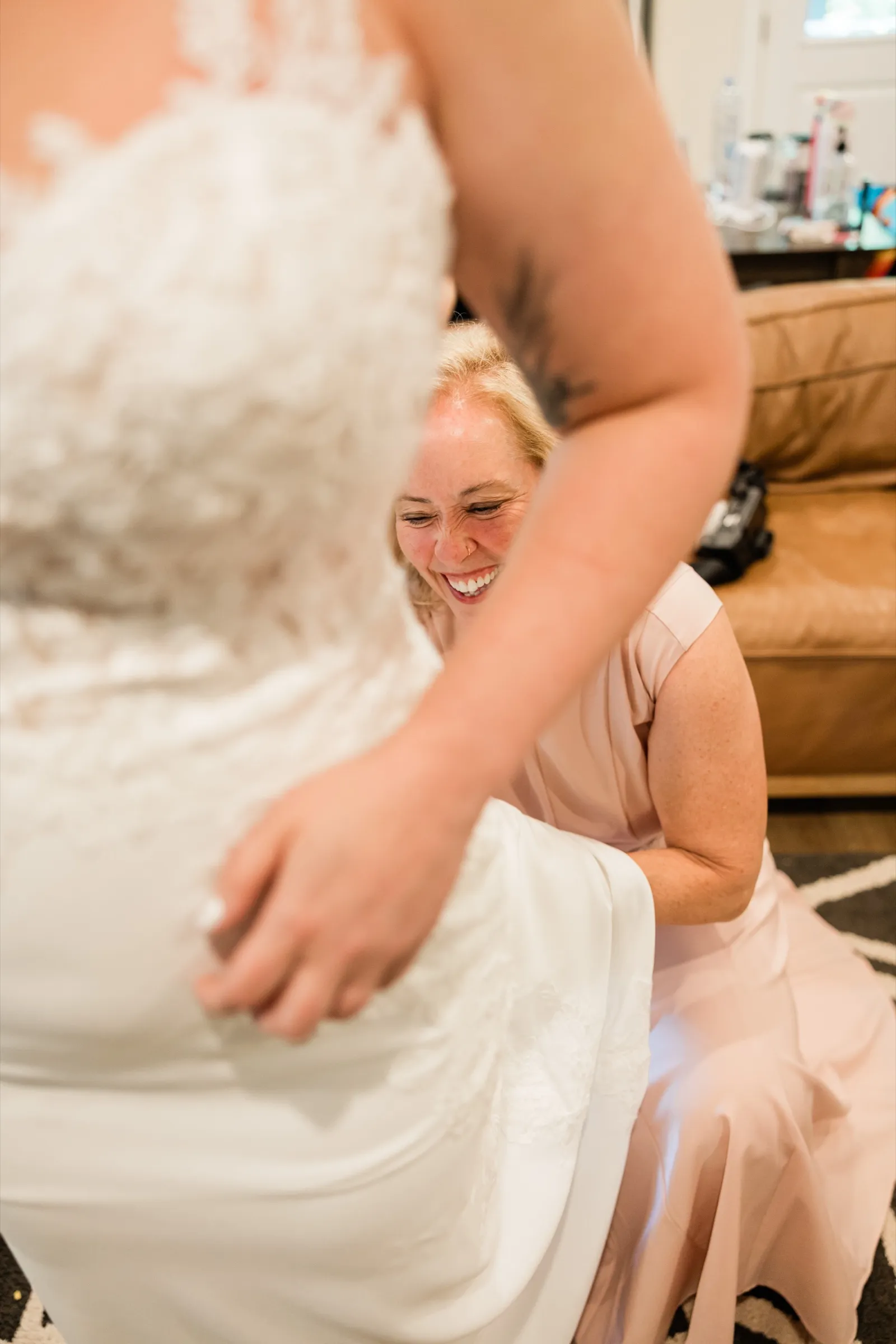A helper in a blush dress laughs while crouched behind the bride, fastening the back of her lace gown — Tim Larsen Photography, Brainerd Lakes MN