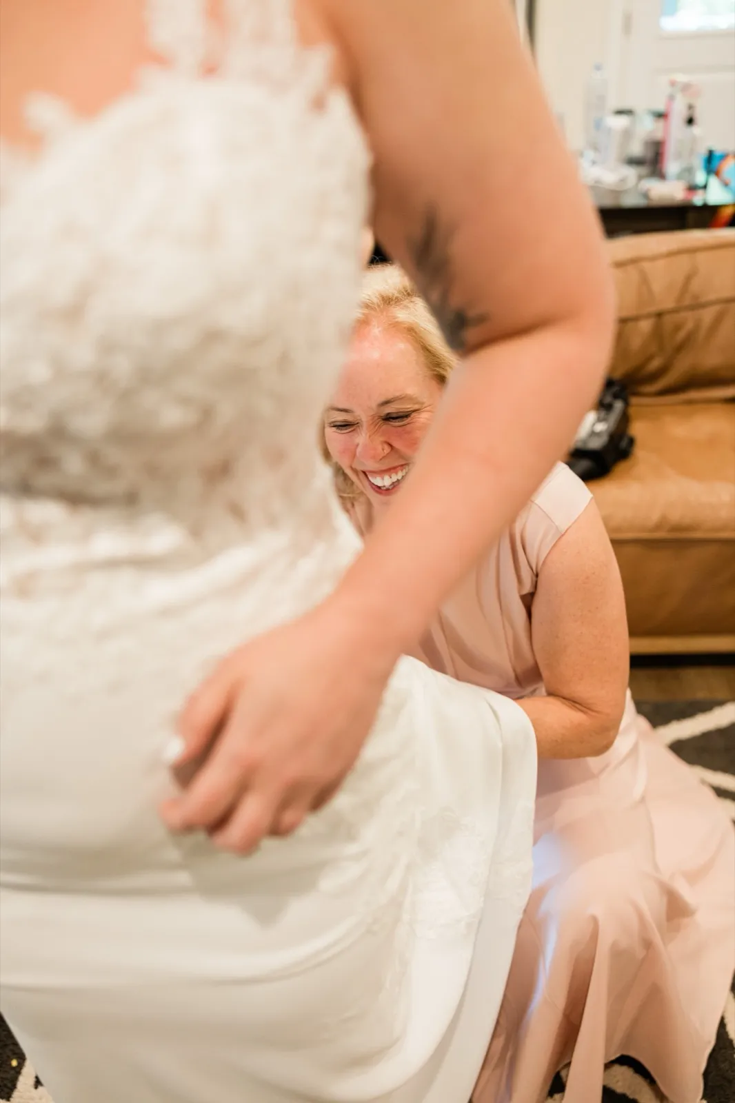 A helper in a blush dress laughs while crouched behind the bride, fastening the back of her lace gown — Tim Larsen Photography, Brainerd Lakes MN