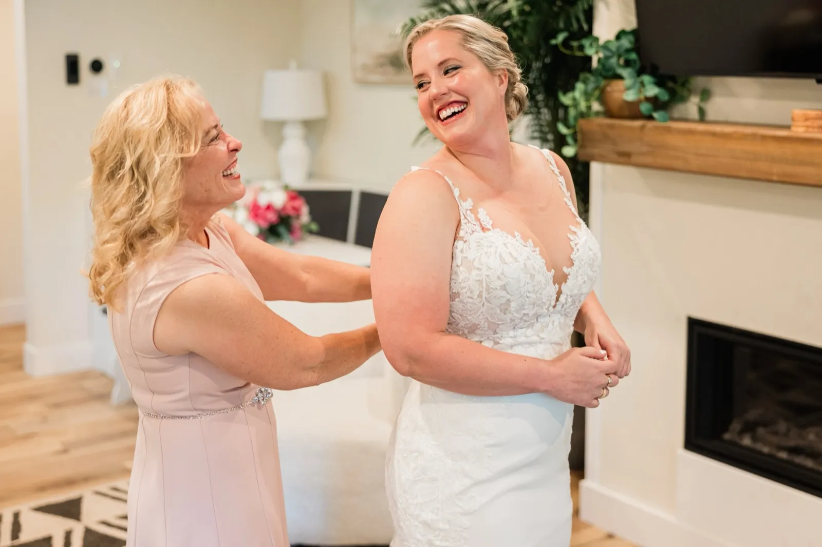 Lauren and her mother laugh together as her mother fastens the back of the lace gown near a fireplace — Tim Larsen Photography, Brainerd Lakes MN
