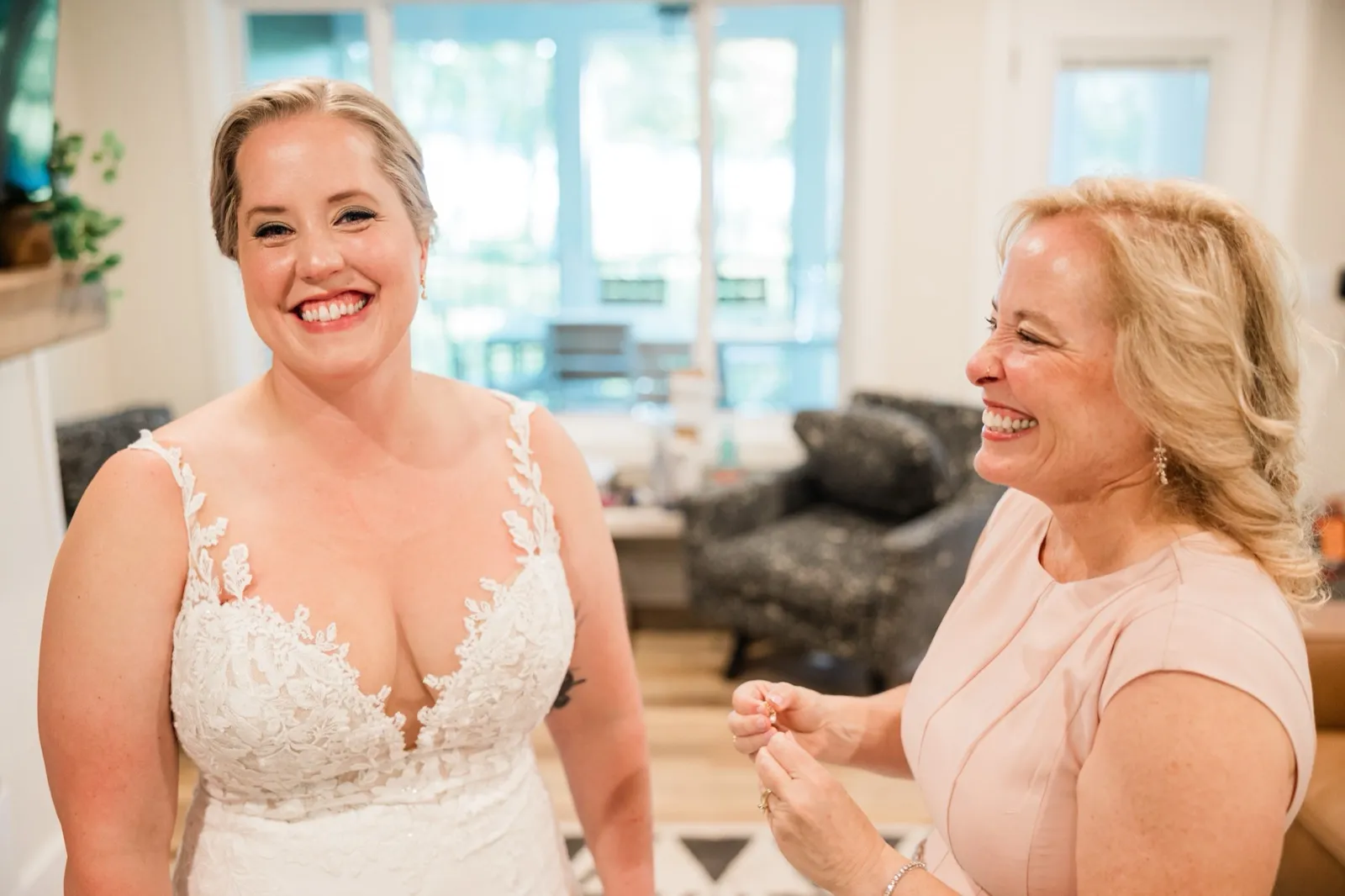 The bride laughs toward the camera while her mother, holding an earring, laughs beside her in a window-lit room — Tim Larsen Photography, Brainerd Lakes MN