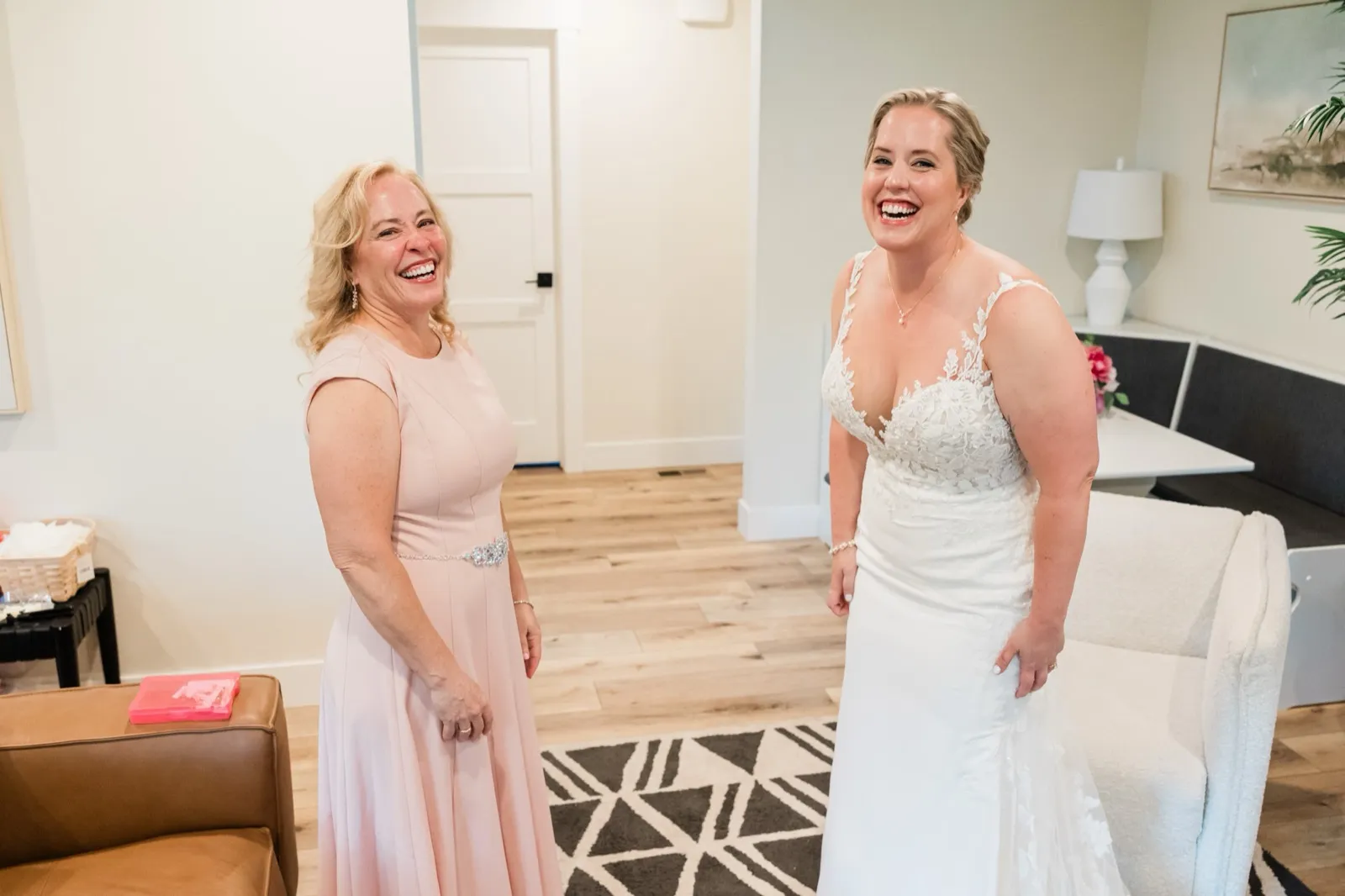 Bride and mother stand laughing together in a bright wood-floored living room on the morning of the wedding — Tim Larsen Photography, Brainerd Lakes MN
