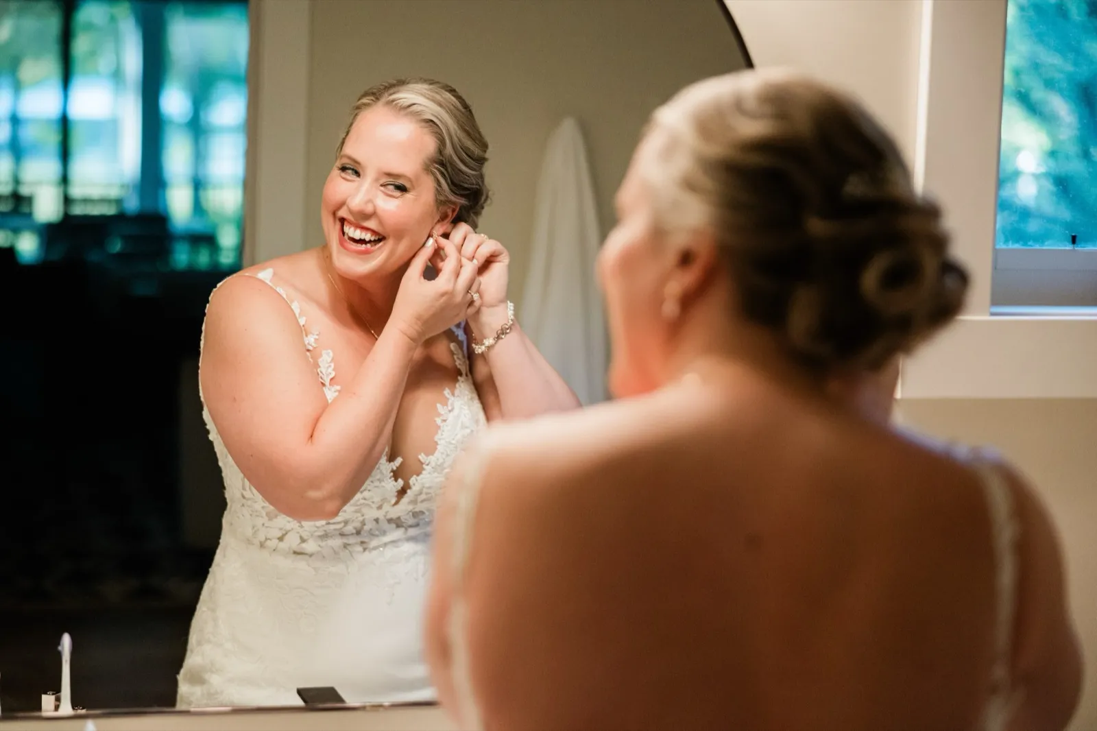 The bride laughs while fastening an earring, her face reflected in a mirror over her bare shoulder — Tim Larsen Photography, Brainerd Lakes MN