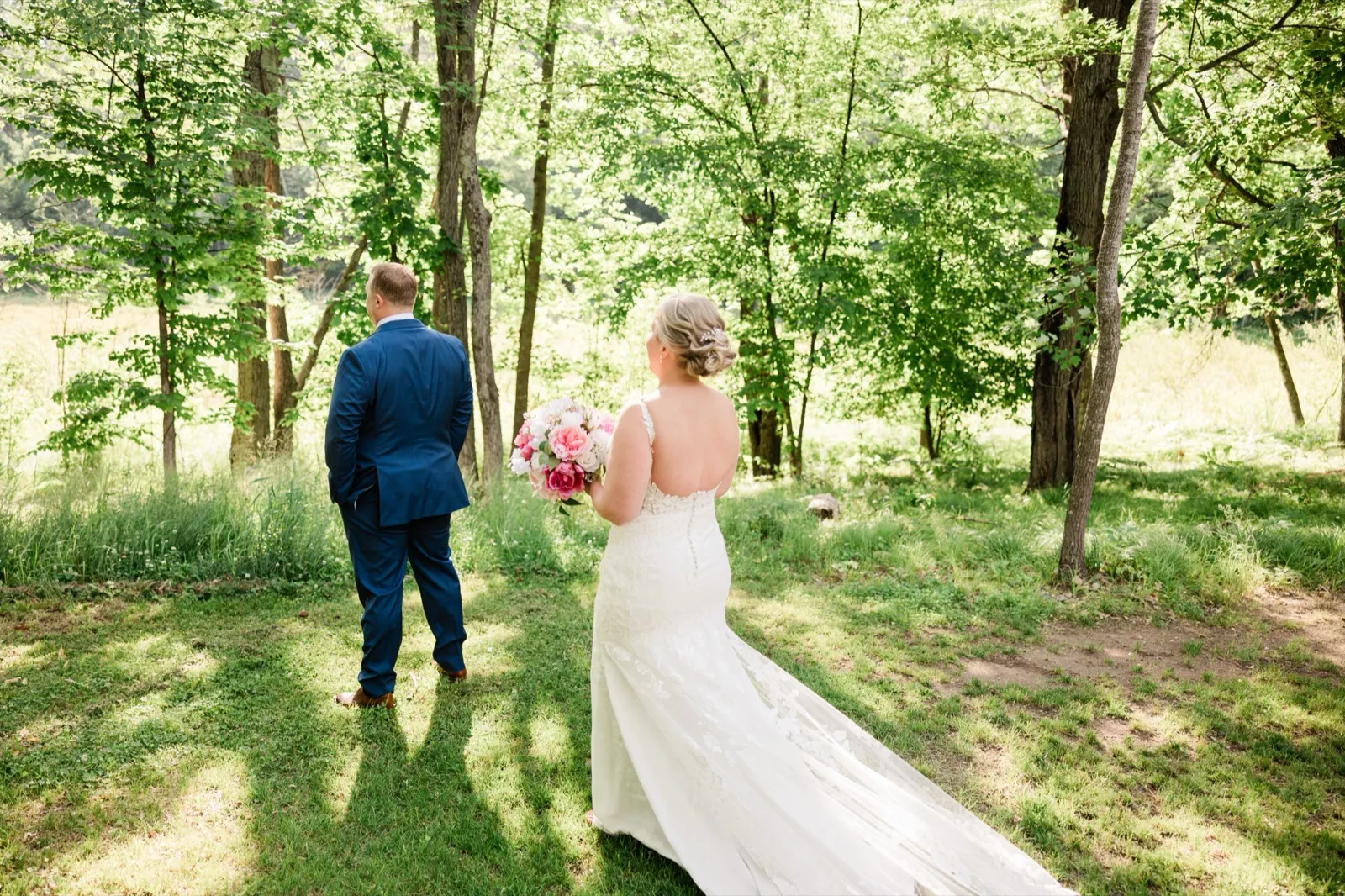Jeremy stands with his back turned in the sunlit woods as Lauren approaches him for the first look — Tim Larsen Photography, Brainerd Lakes MN