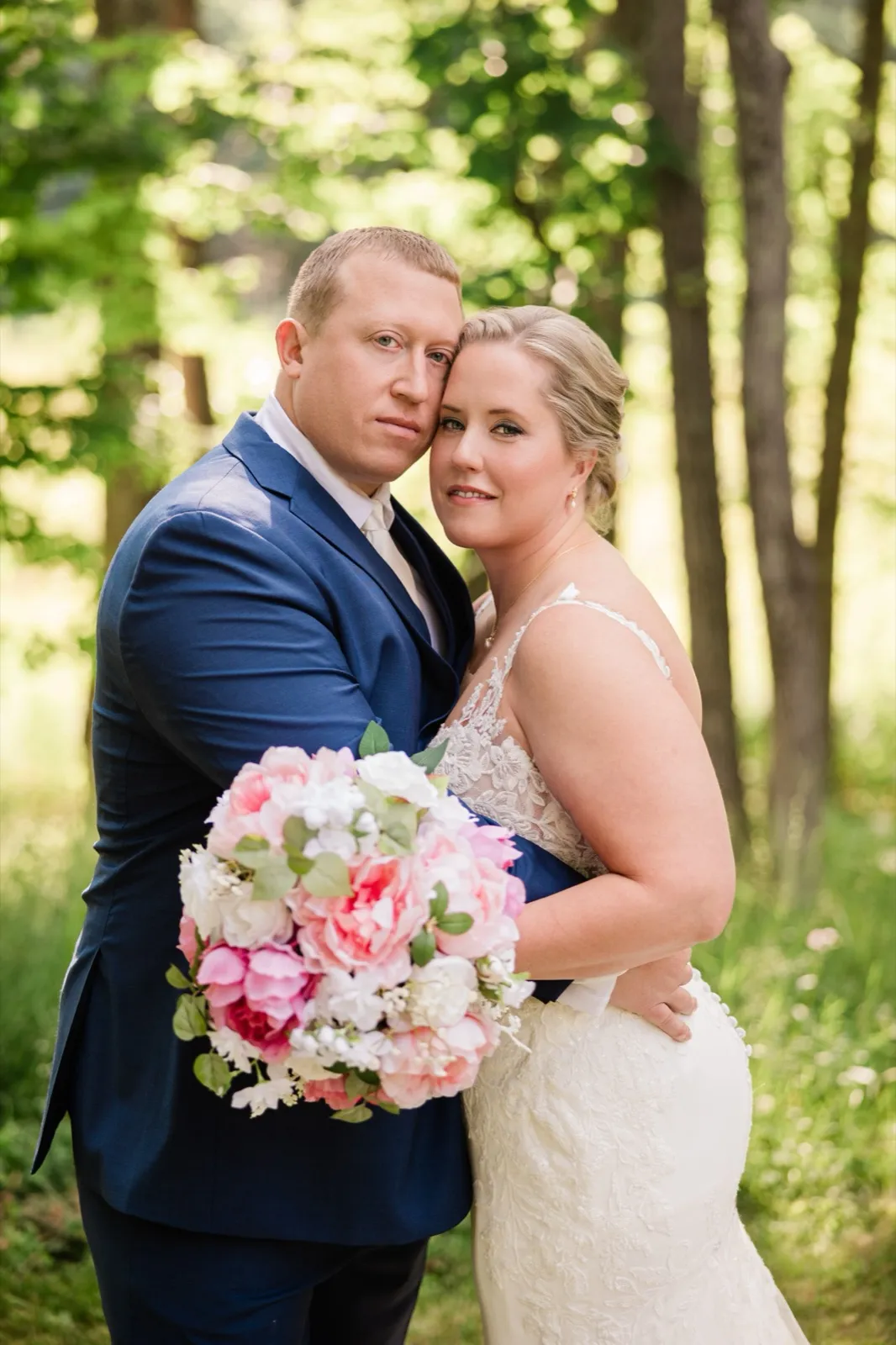 The couple stands cheek to cheek in the sunlit forest, the bride holding her pink bouquet — Tim Larsen Photography, Brainerd Lakes MN
