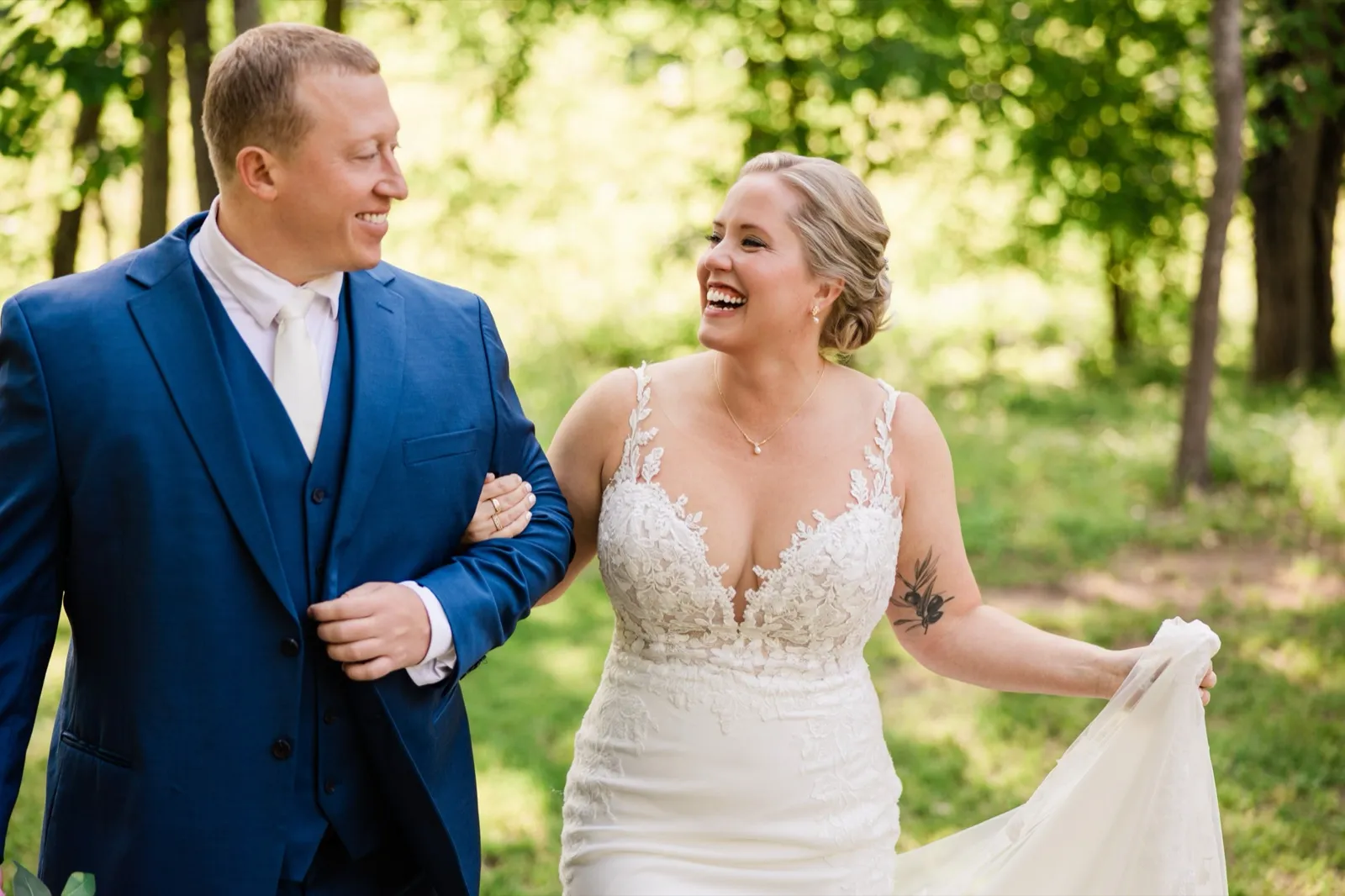 The bride laughs wide open walking arm-in-arm with the groom, lifting her train off the woodland path — Tim Larsen Photography, Brainerd Lakes MN