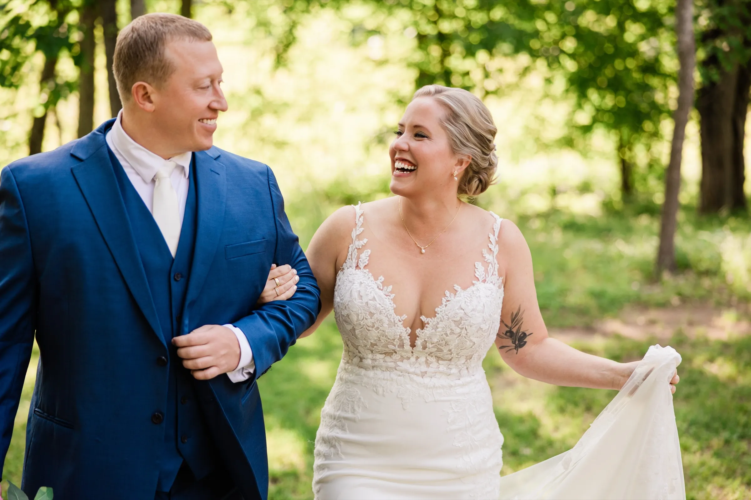 The bride laughs wide open walking arm-in-arm with the groom, lifting her train off the woodland path — Tim Larsen Photography, Brainerd Lakes MN