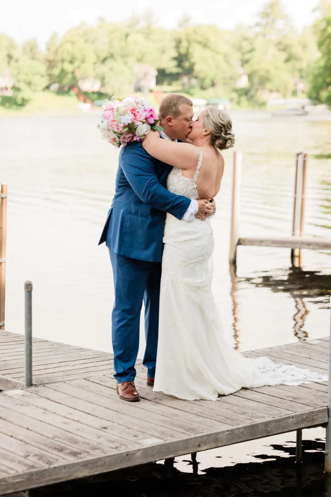 The couple kisses at the edge of a wooden dock over the lake, her bouquet wrapped behind his neck — Tim Larsen Photography, Brainerd Lakes MN