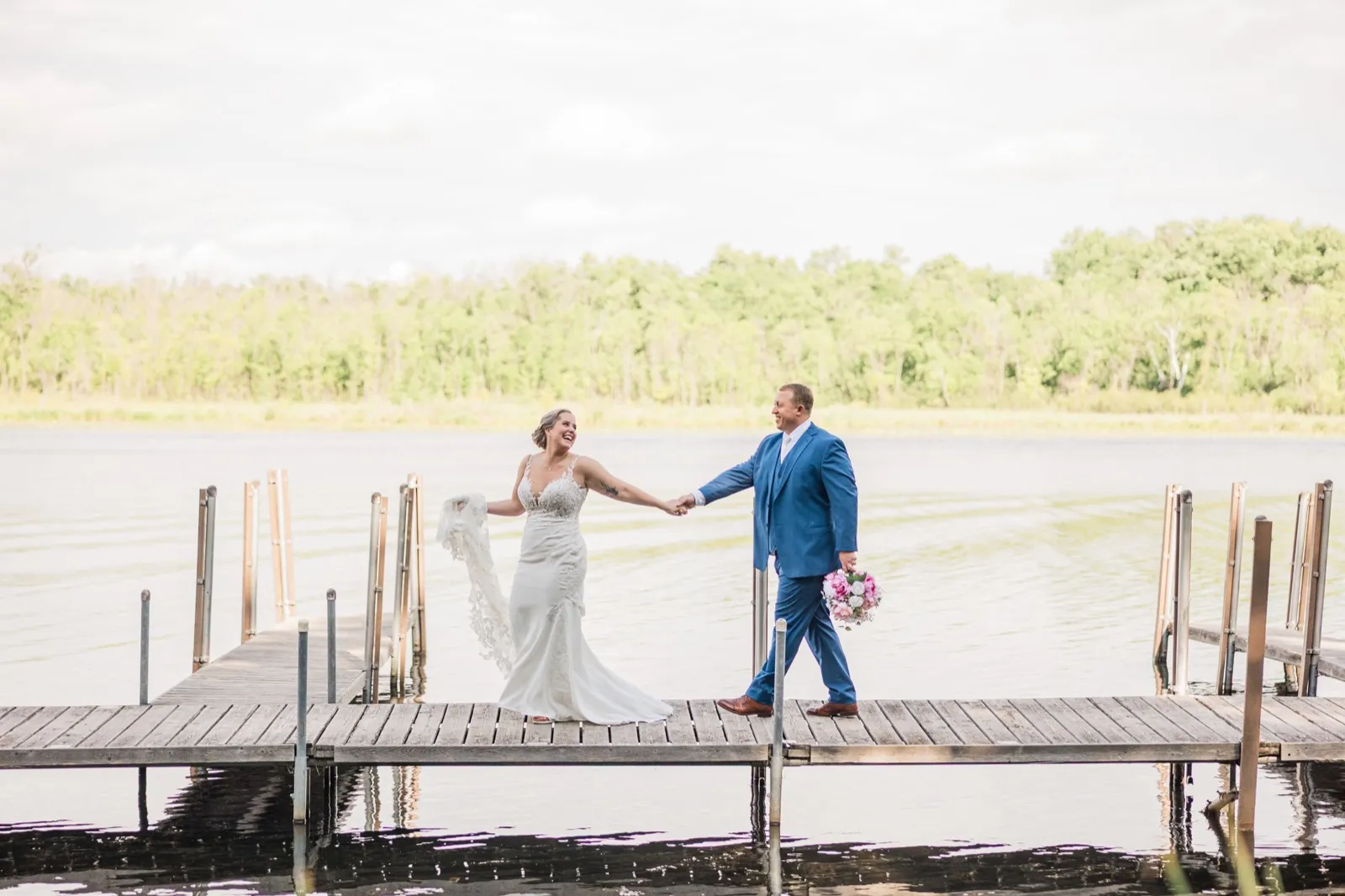 Jeremy leads Lauren by the hand across the dock at Lost Lake Lodge, carrying her bouquet as she laughs and holds her veil — Tim Larsen Photography, Brainerd Lakes MN