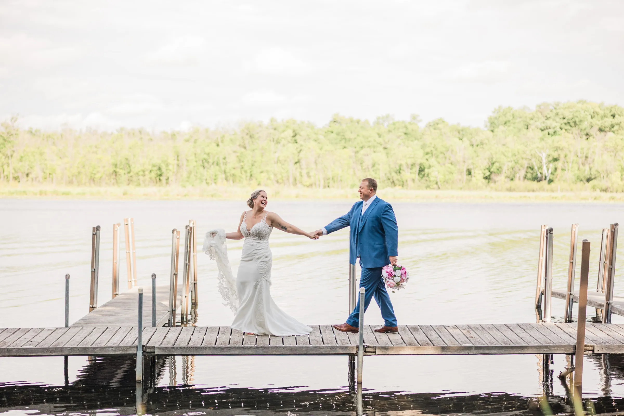 Jeremy leads Lauren by the hand across the dock at Lost Lake Lodge, carrying her bouquet as she laughs and holds her veil — Tim Larsen Photography, Brainerd Lakes MN