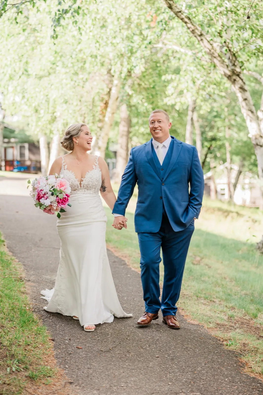 The couple walks hand in hand down a tree-lined path, Lauren laughing with her bright pink bouquet — Tim Larsen Photography, Brainerd Lakes MN