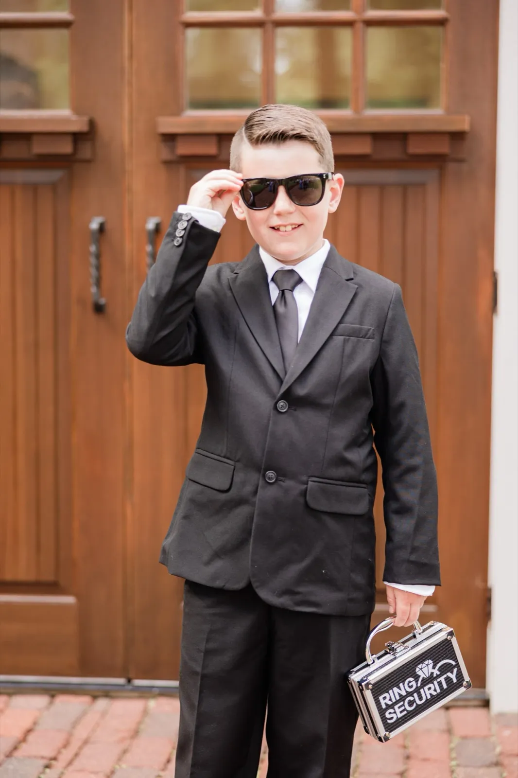 The young ring bearer tips his sunglasses down and grins, holding a case labeled 'Ring Security' outside the lodge doors — Tim Larsen Photography, Brainerd Lakes MN