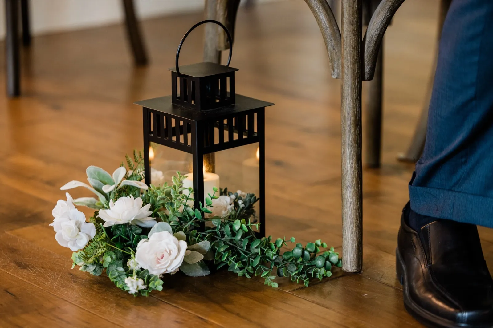 A black lantern with a lit candle sits beside white roses and eucalyptus along the chapel aisle — Tim Larsen Photography, Brainerd Lakes MN