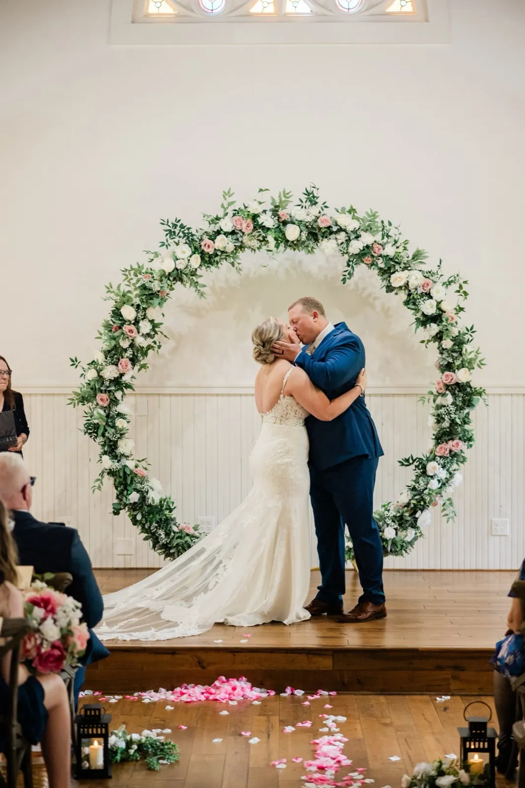 Lauren and Jeremy share their first kiss centered in the circular floral arch, stained glass glowing above — Tim Larsen Photography, Brainerd Lakes MN