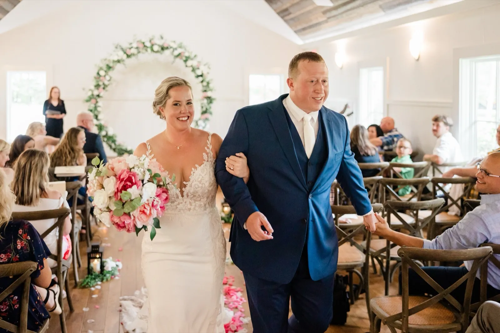 The beaming newlyweds walk back up the aisle as Jeremy reaches to clasp a seated guest's hand — Tim Larsen Photography, Brainerd Lakes MN