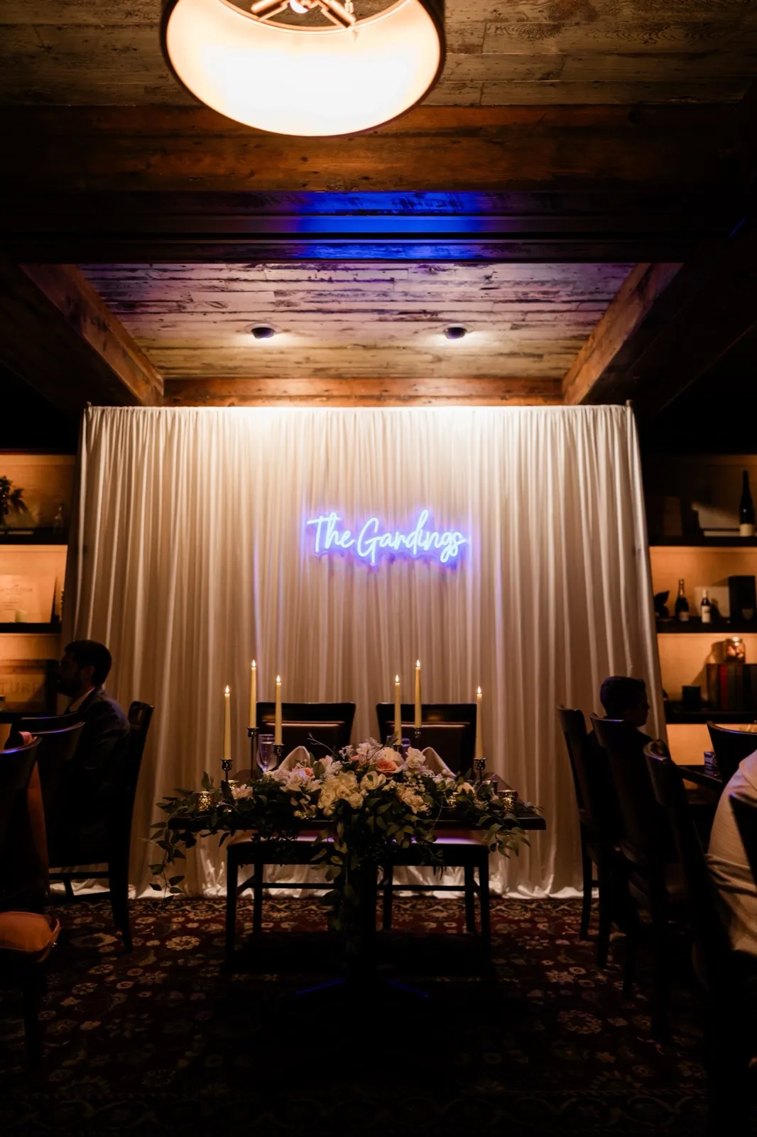 The candlelit sweetheart table under the glowing neon 'The Gardings' sign at Cru, taper candles and garden roses waiting for the newlyweds — Tim Larsen Photography, Brainerd Lakes MN