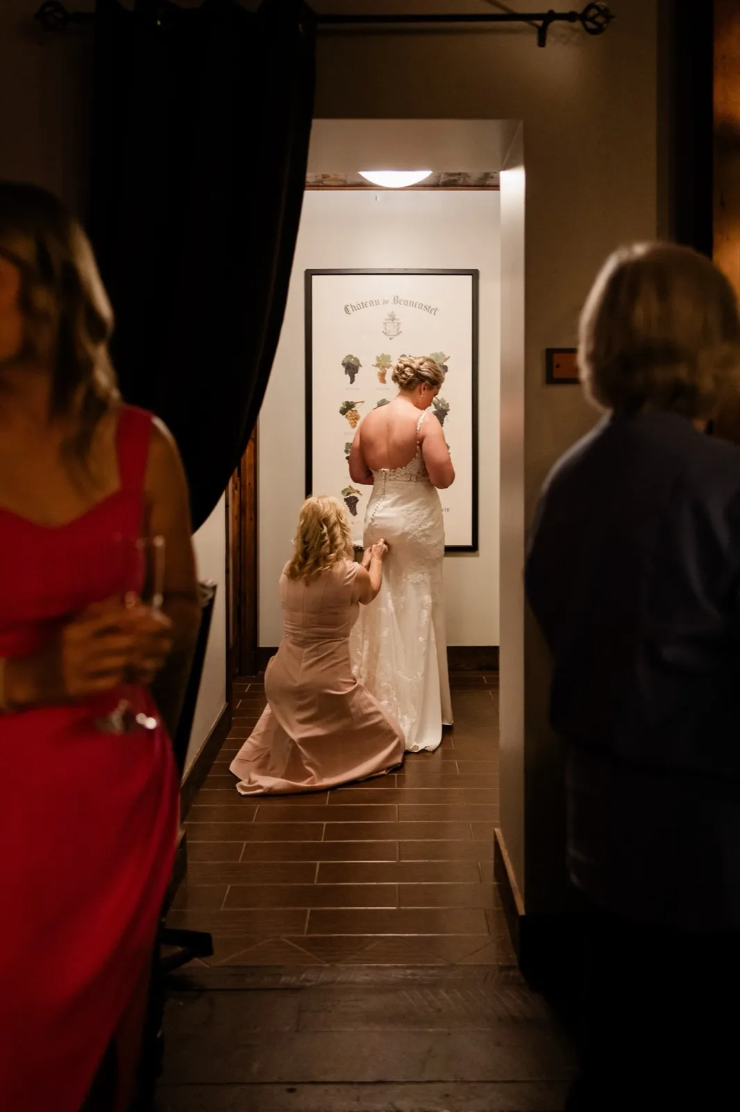Framed through a doorway, Lauren's mom kneels in her blush dress to fasten the bustle in the hallway at Cru — Tim Larsen Photography, Brainerd Lakes MN