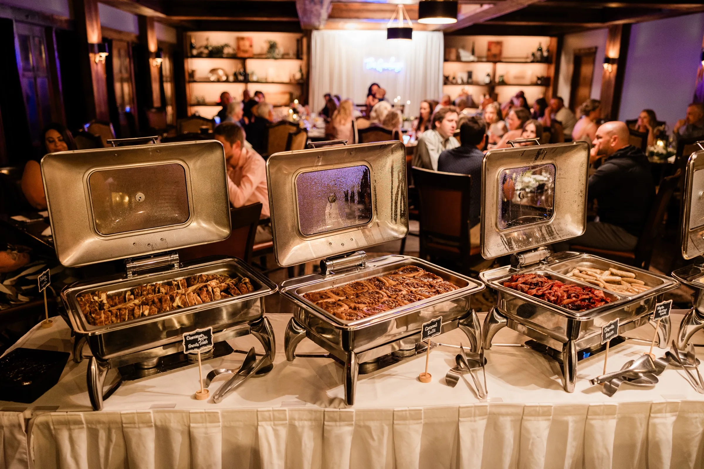 Chafing dishes of french toast, caramel rolls, bacon, and sausage line the brunch buffet as guests dine behind — Tim Larsen Photography, Brainerd Lakes MN