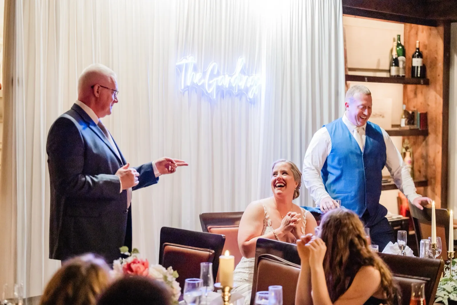 Lauren's father points mid-toast as she laughs and claps and Jeremy stands laughing beside her under the neon 'The Gardings' sign — Tim Larsen Photography, Brainerd Lakes MN