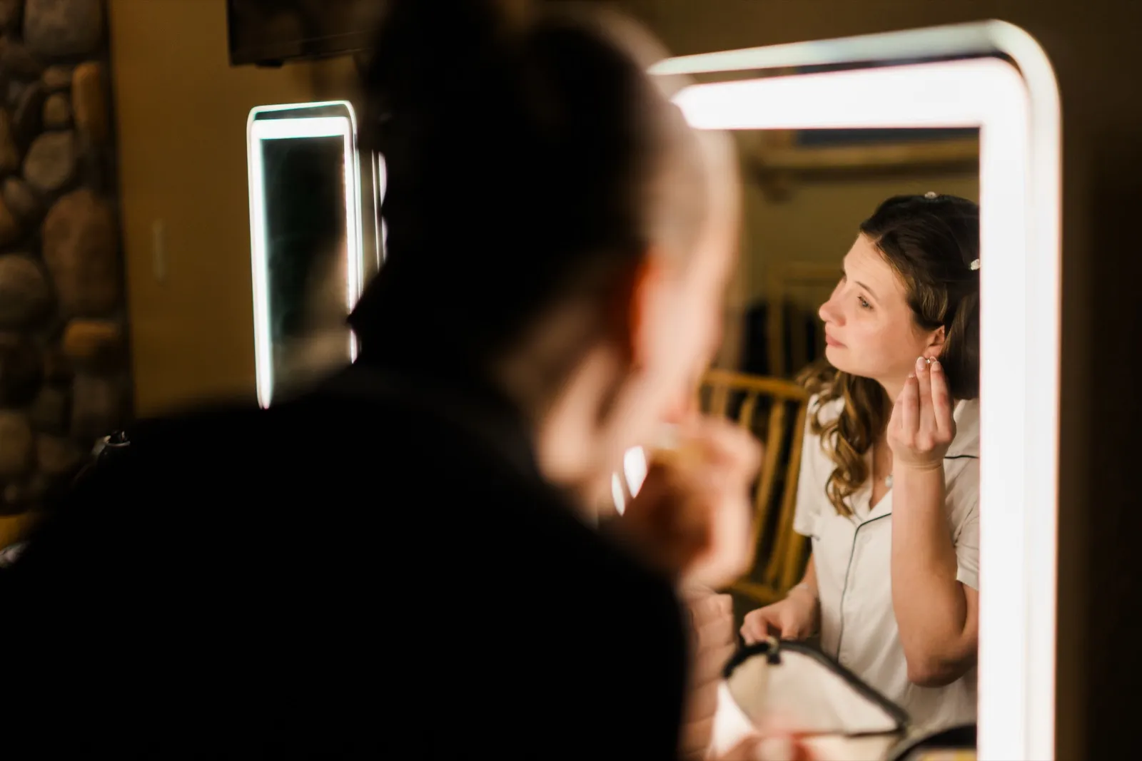 Maddie fastens an earring while looking into a lighted vanity mirror — Tim Larsen Photography, Brainerd Lakes MN