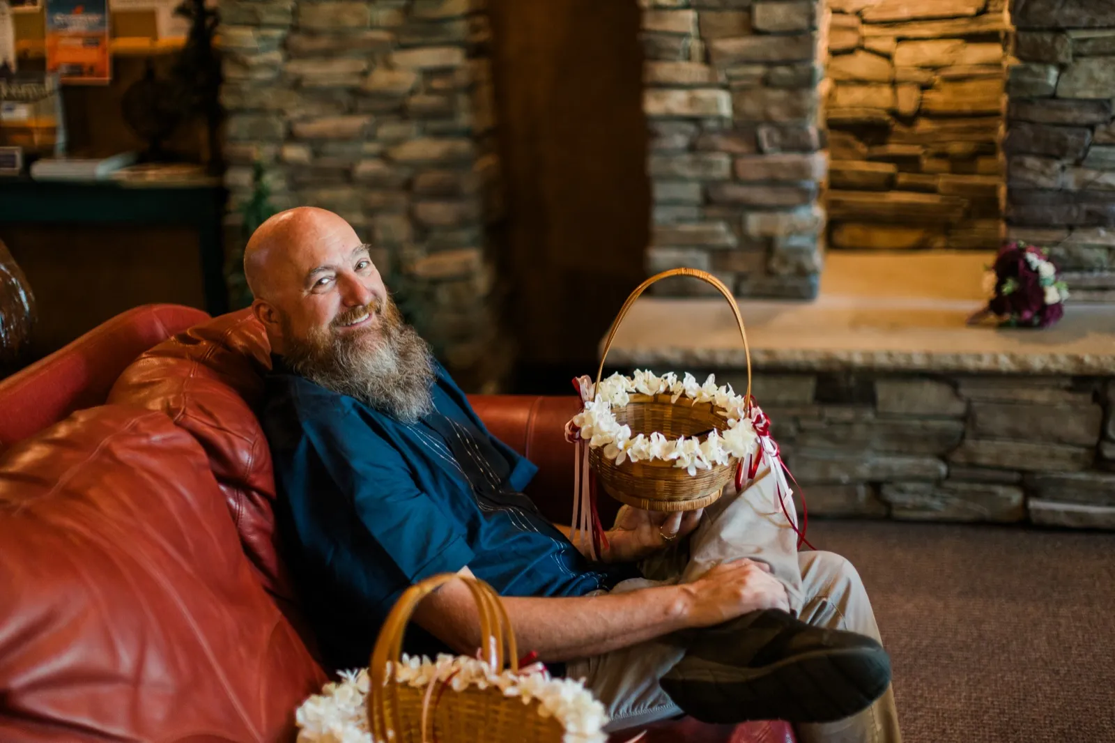 A bearded man sits on a red leather couch holding flower baskets beside the lodge fireplace — Tim Larsen Photography, Brainerd Lakes MN