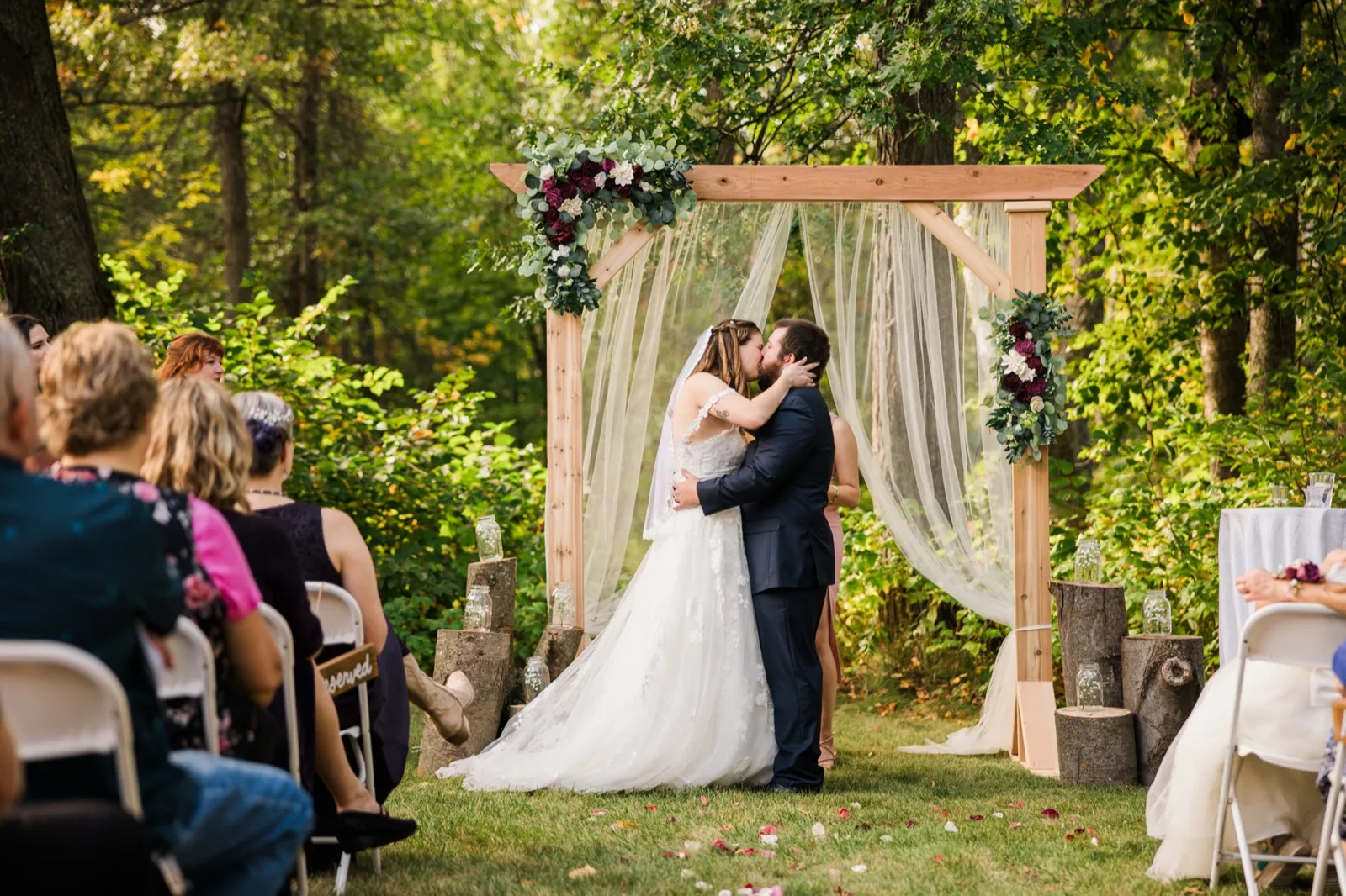 Maddie and Cody share their ceremony kiss beneath a wooden arch with fabric and burgundy flowers — Tim Larsen Photography, Brainerd Lakes MN