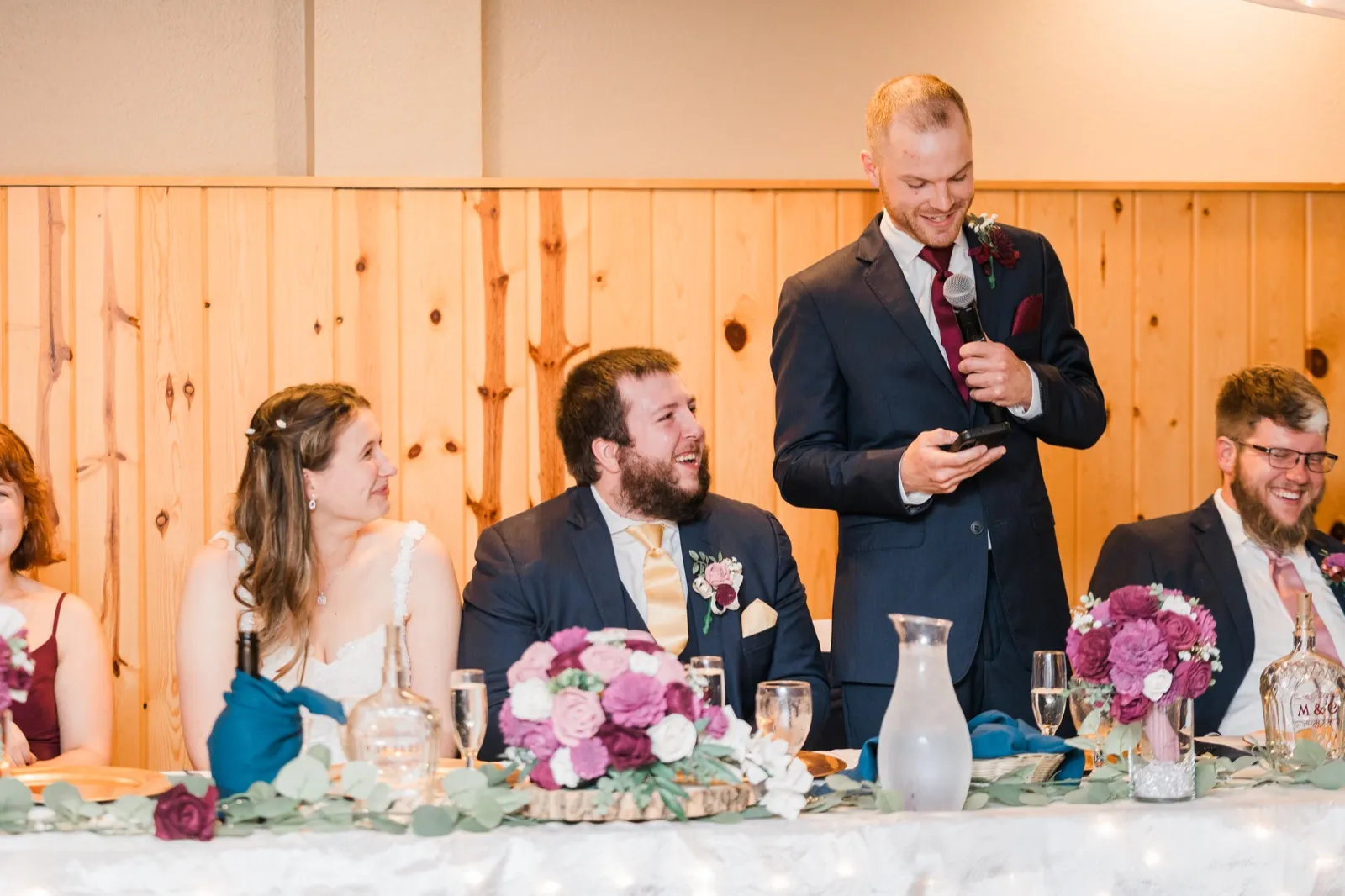 A groomsman reads a toast from his phone while Maddie and Cody listen at the head table — Tim Larsen Photography, Brainerd Lakes MN