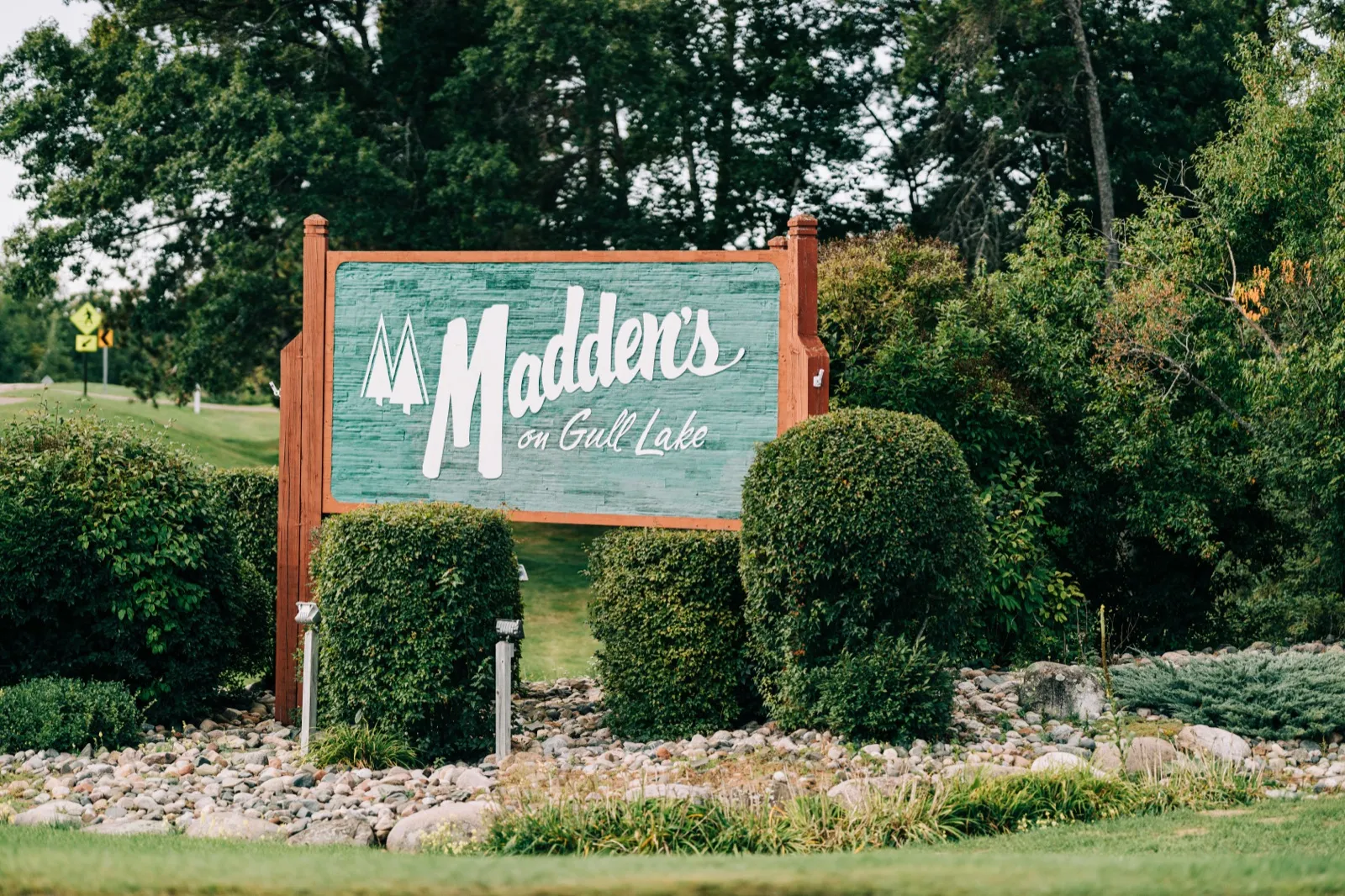 The painted teal entrance sign for Madden's on Gull Lake stands among manicured hedges and tall trees on an overcast afternoon. — Tim Larsen Photography, Brainerd Lakes MN