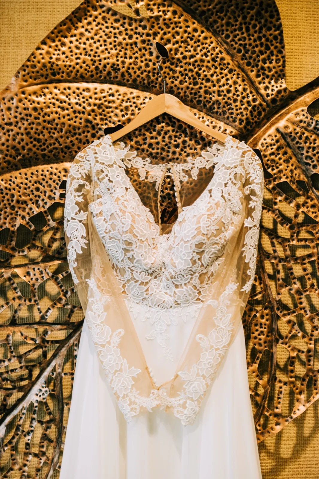 A lace wedding gown with a sheer floral bodice hangs from a wooden hanger against an ornate copper-toned leaf-shaped wall sculpture. — Tim Larsen Photography, Brainerd Lakes MN