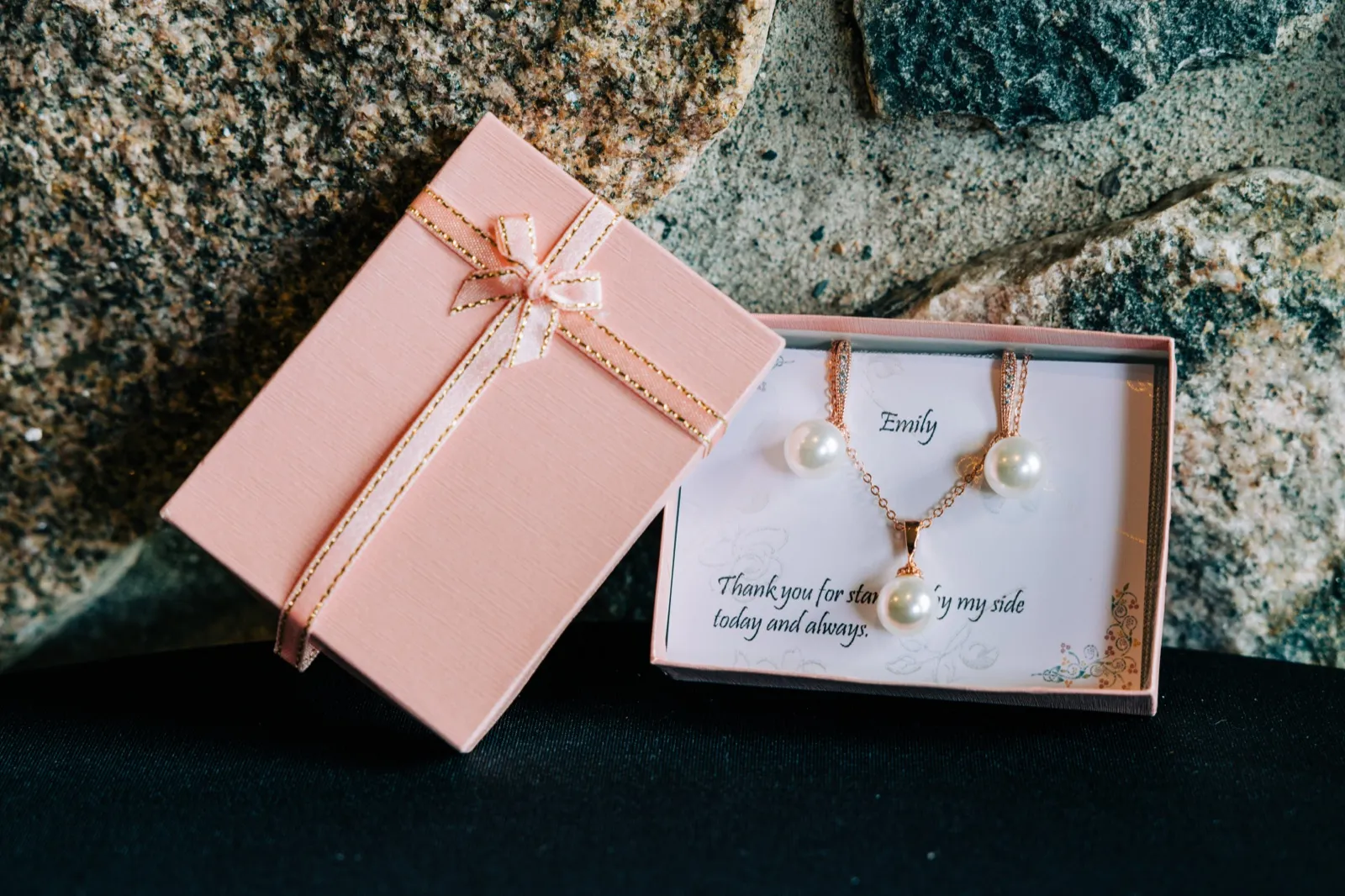 A pink gift box with a ribbon bow leans against an open box containing a pearl necklace and earrings on a card reading 'Emily, thank you for standing by my side today and always,' set against a textured stone backdrop. — Tim Larsen Photography, Brainerd Lakes MN