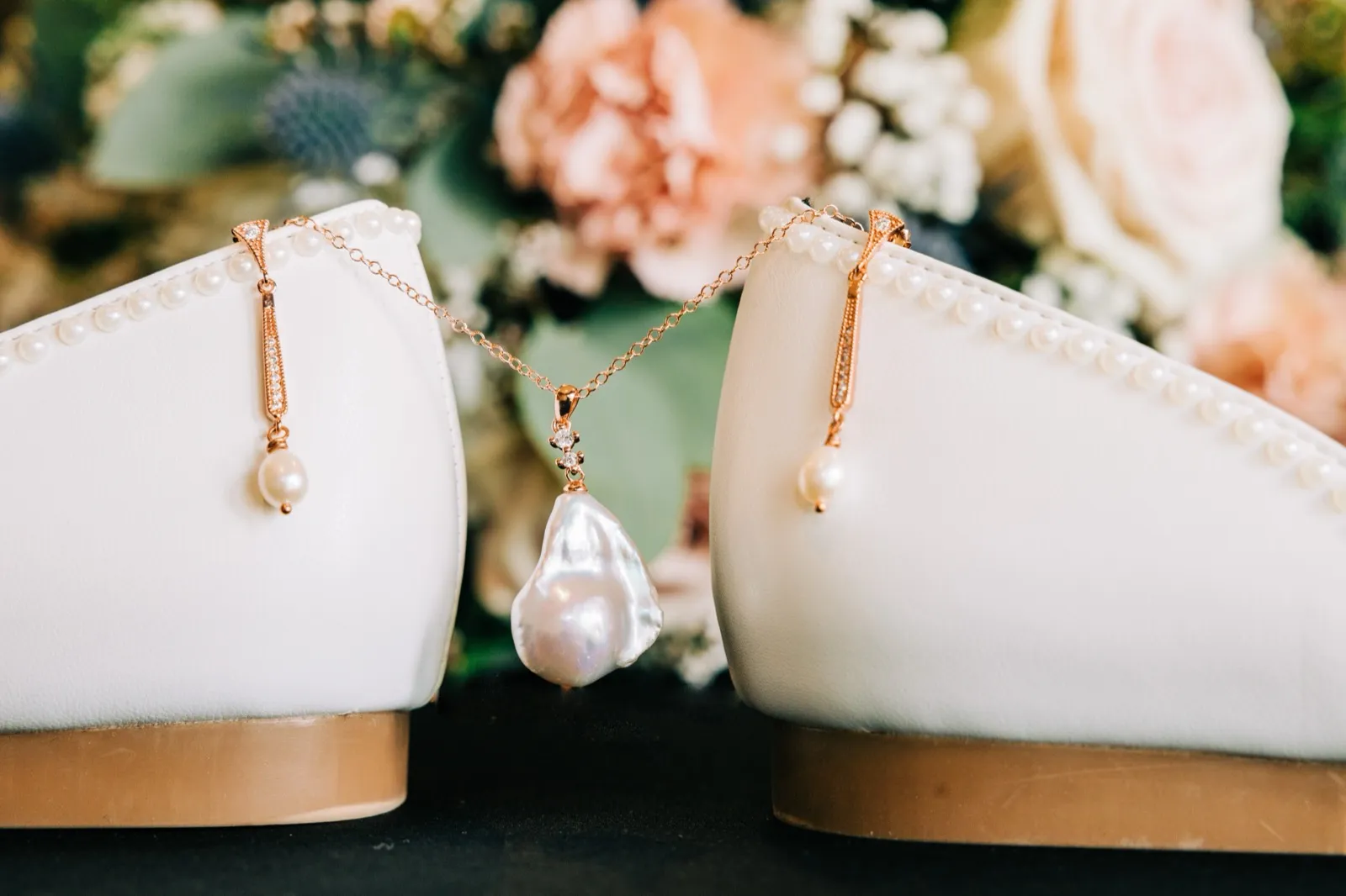 A baroque pearl pendant necklace drapes between a pair of pearl-trimmed ivory bridal flats, with a blush and white bouquet softly blurred behind. — Tim Larsen Photography, Brainerd Lakes MN