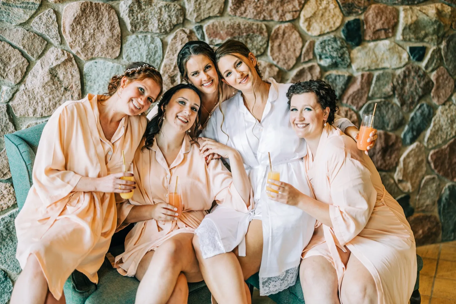 The bride and four bridesmaids in matching peach robes sit close together holding mimosas in front of a large stone wall. — Tim Larsen Photography, Brainerd Lakes MN