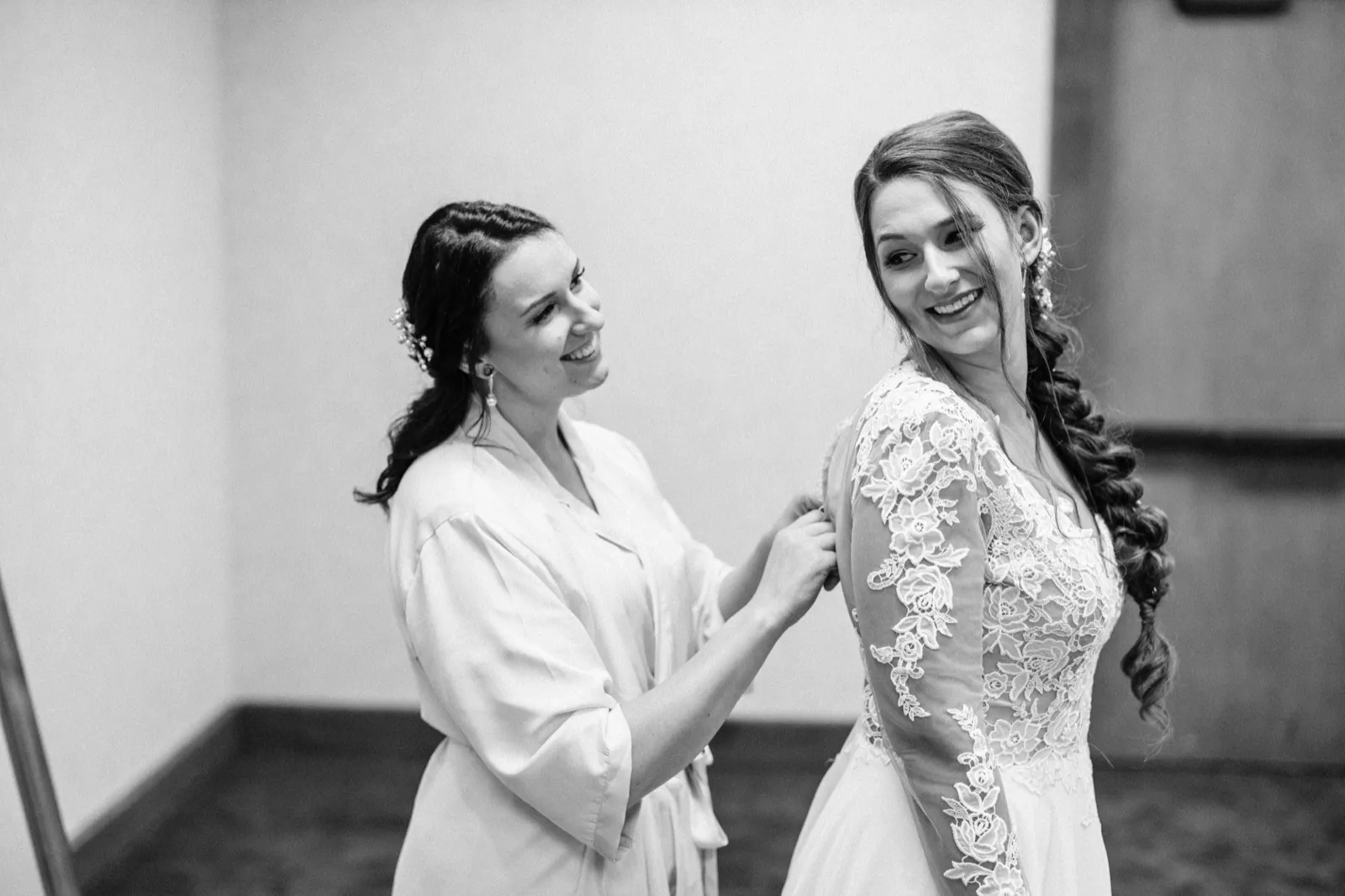 In black and white, a bridesmaid in a satin robe smiles at the bride while buttoning the back of her lace wedding gown. — Tim Larsen Photography, Brainerd Lakes MN