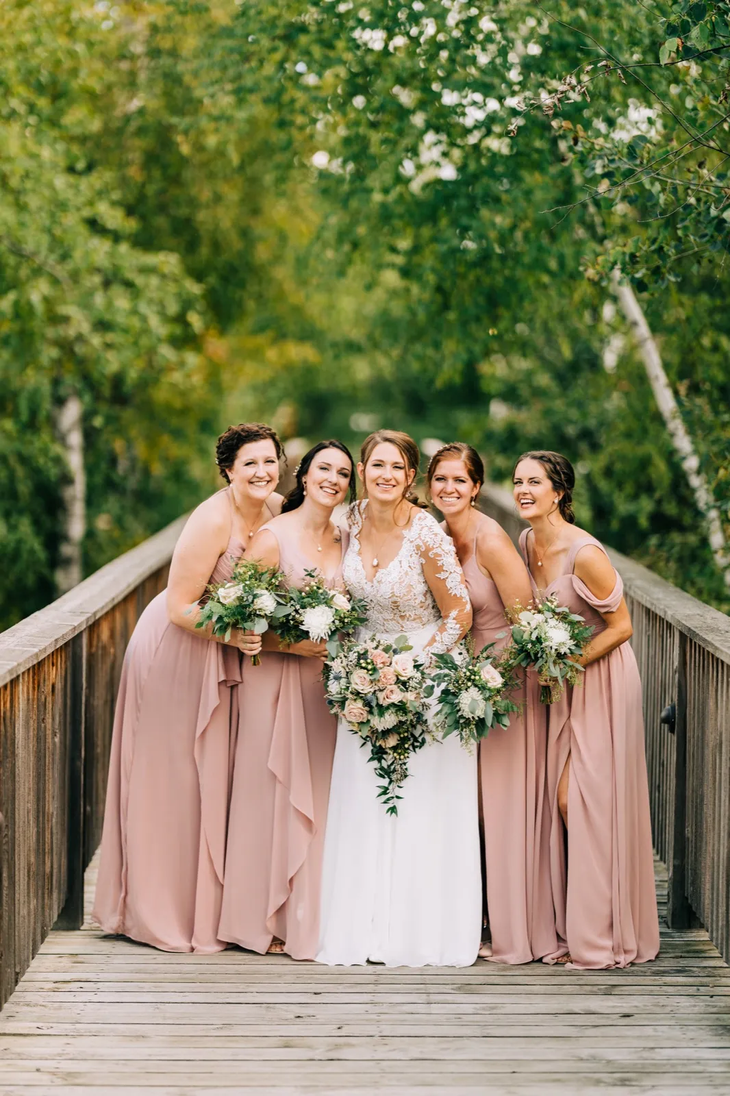 The bride stands centered with four bridesmaids in dusty pink dresses on a wooden footbridge, all laughing while holding cascading bouquets. — Tim Larsen Photography, Brainerd Lakes MN