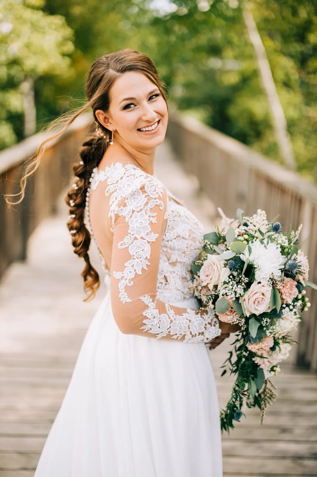 The bride turns to look over her shoulder on a wooden footbridge, smiling broadly while holding a cascading bouquet of blush roses, white blooms, and blue thistle. — Tim Larsen Photography, Brainerd Lakes MN