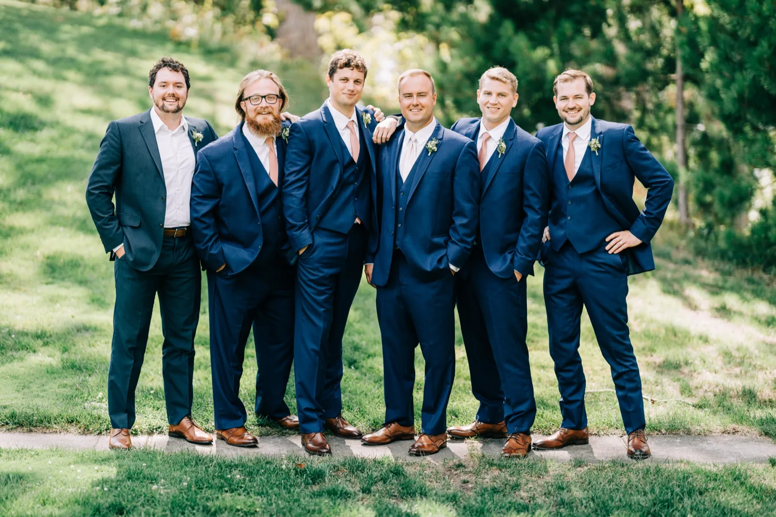 The groom stands with five groomsmen in navy suits and blush ties on a grassy slope, smiling toward the camera. — Tim Larsen Photography, Brainerd Lakes MN