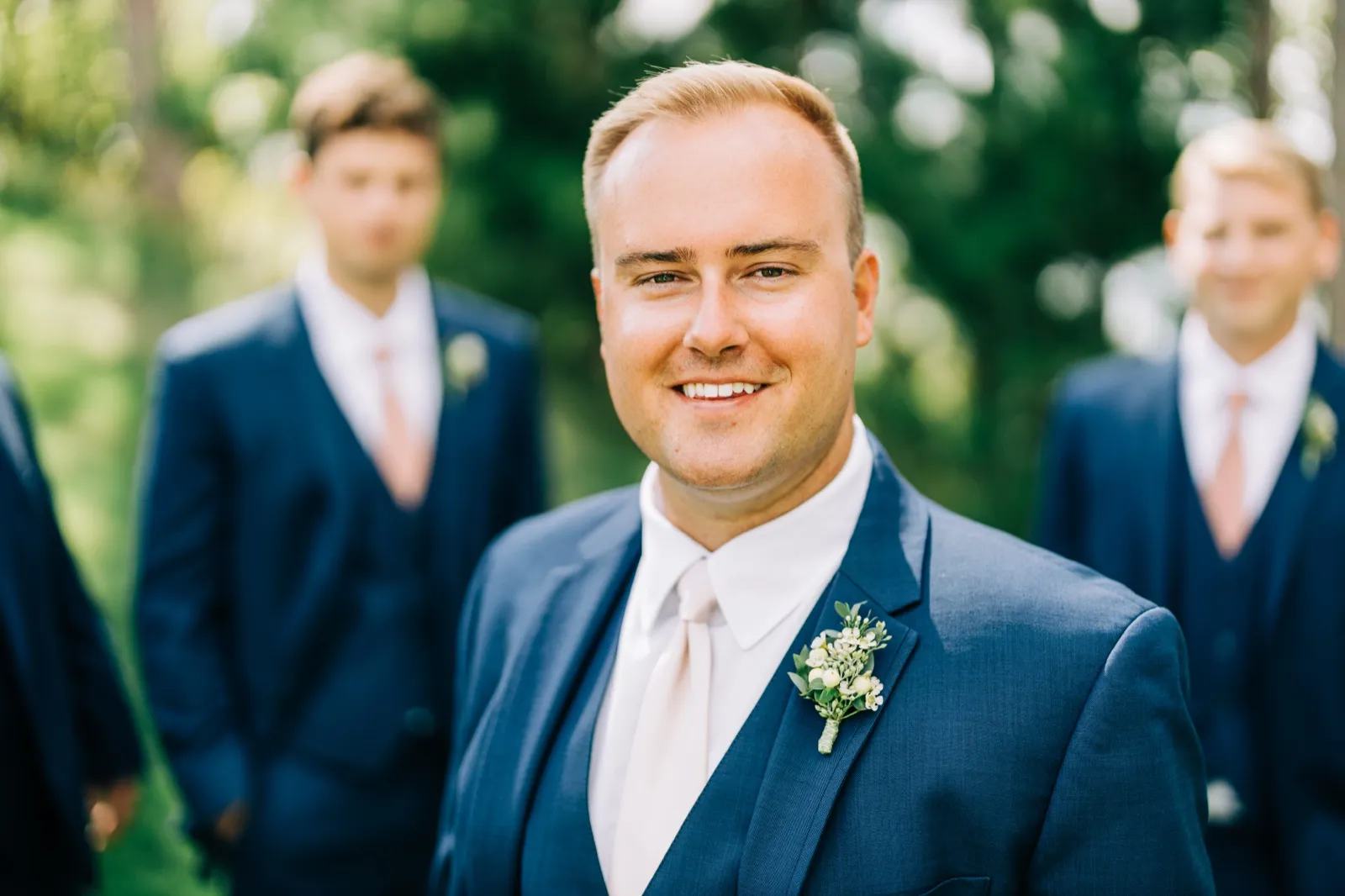 The groom smiles at the camera in a navy suit and white tie, with two groomsmen softly blurred behind him among trees. — Tim Larsen Photography, Brainerd Lakes MN