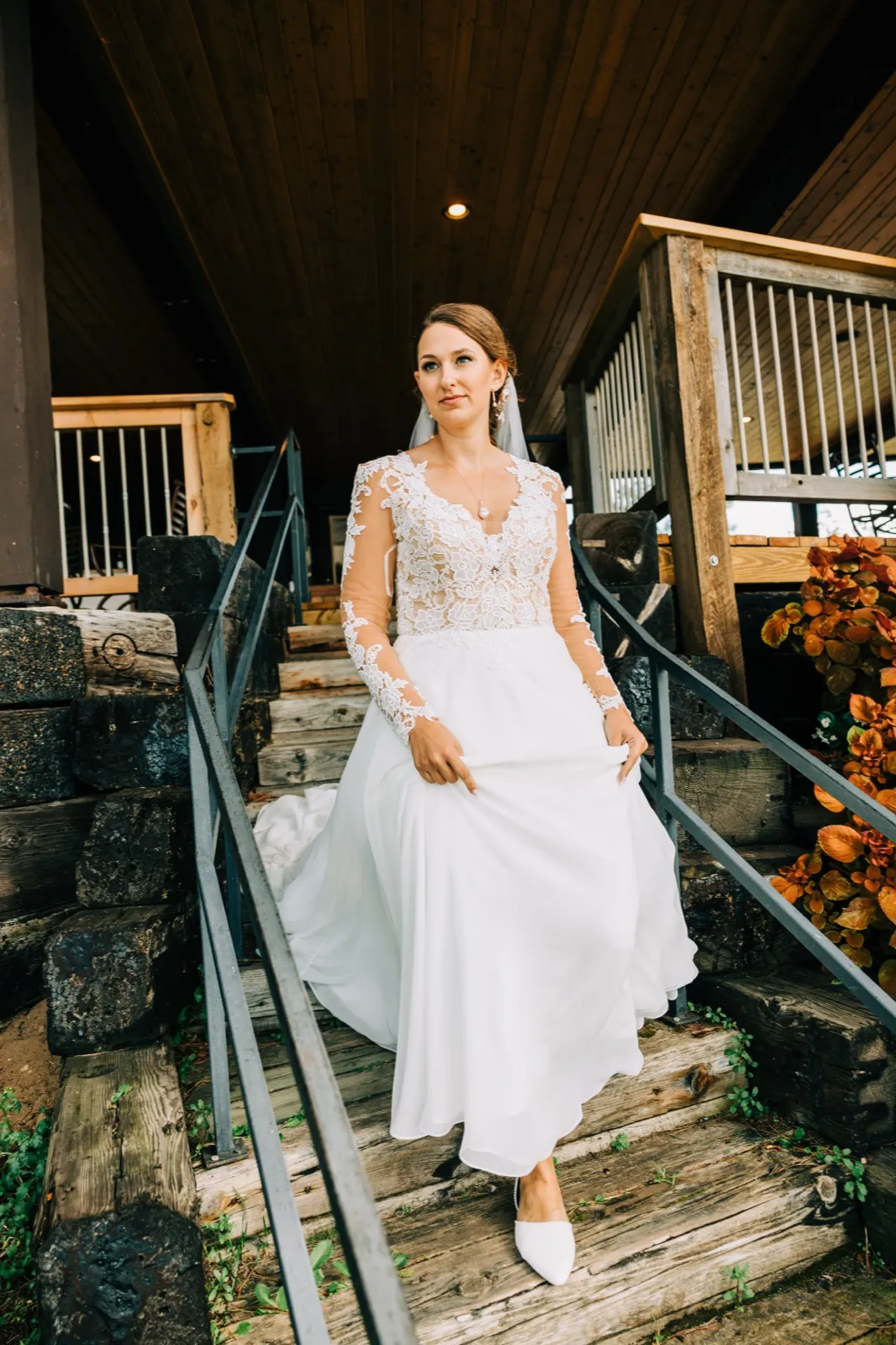 The bride pauses on a rustic wooden staircase in a long-sleeved lace gown and veil, with the A-frame eaves of the lodge above her and autumn foliage to her right. — Tim Larsen Photography, Brainerd Lakes MN