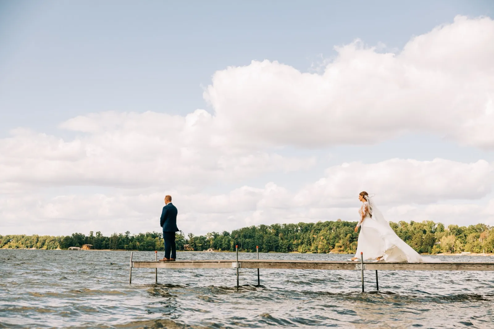 The groom stands waiting at the end of a long lake dock as the bride approaches from the right with her veil and train trailing behind her, Gull Lake stretching out beneath a wide cloud-filled sky. — Tim Larsen Photography, Brainerd Lakes MN