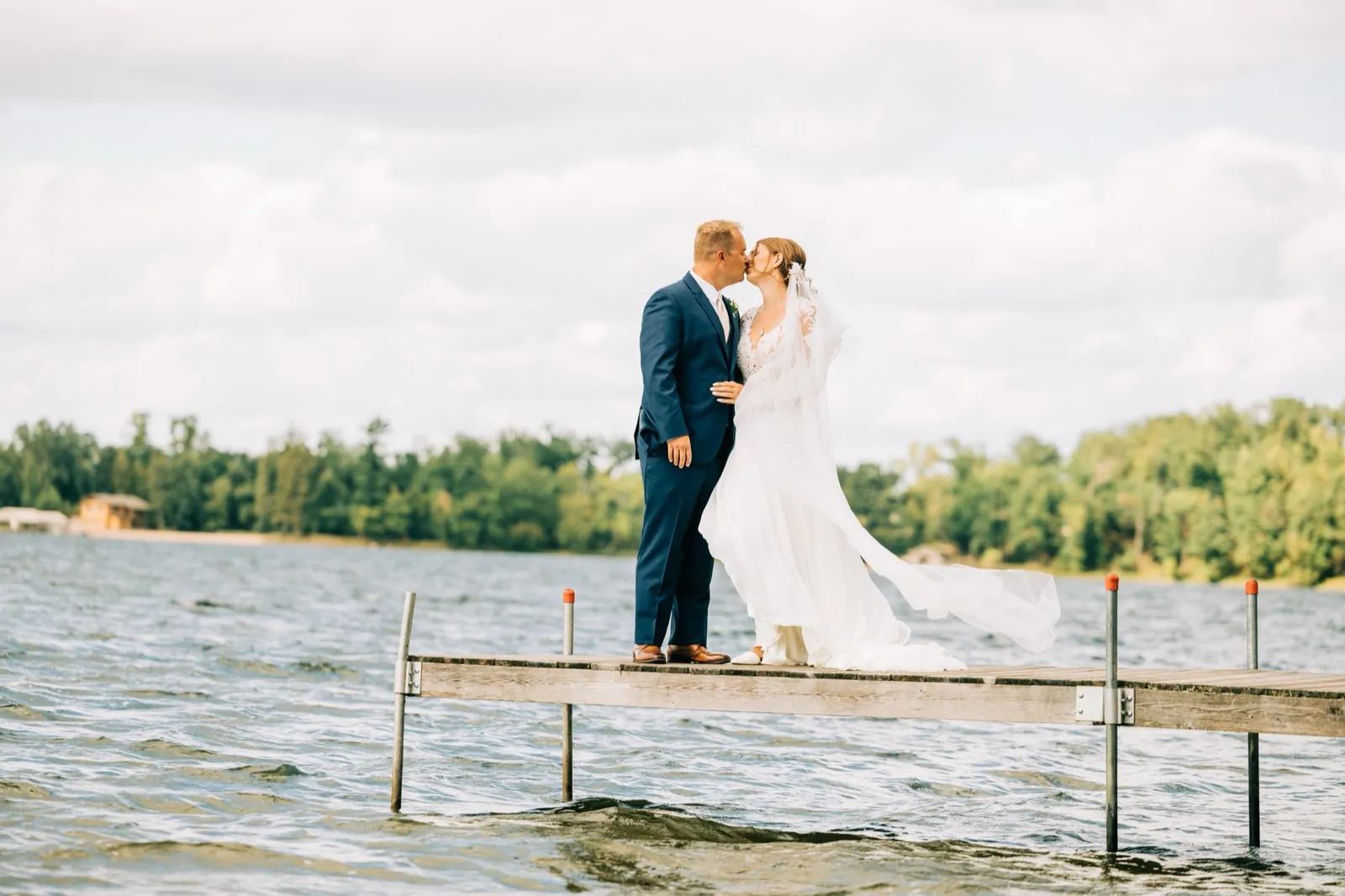 The bride and groom share a kiss on a wooden dock extending into Gull Lake, the bride's long veil trailing sideways in the wind. — Tim Larsen Photography, Brainerd Lakes MN