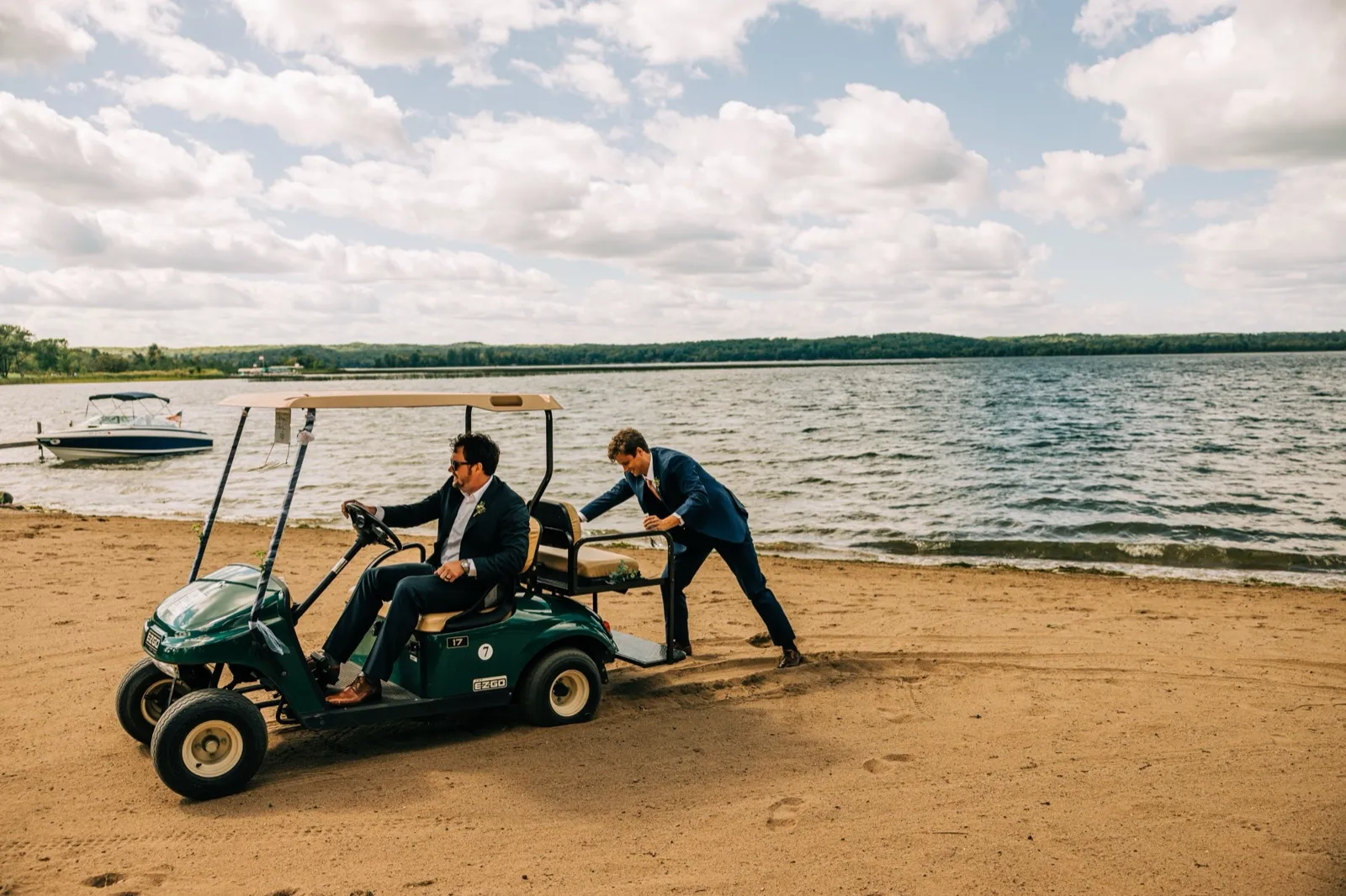 A groomsman in a navy suit pushes a golf cart stuck in beach sand while the groom sits at the wheel, with Gull Lake and a moored boat behind them. — Tim Larsen Photography, Brainerd Lakes MN