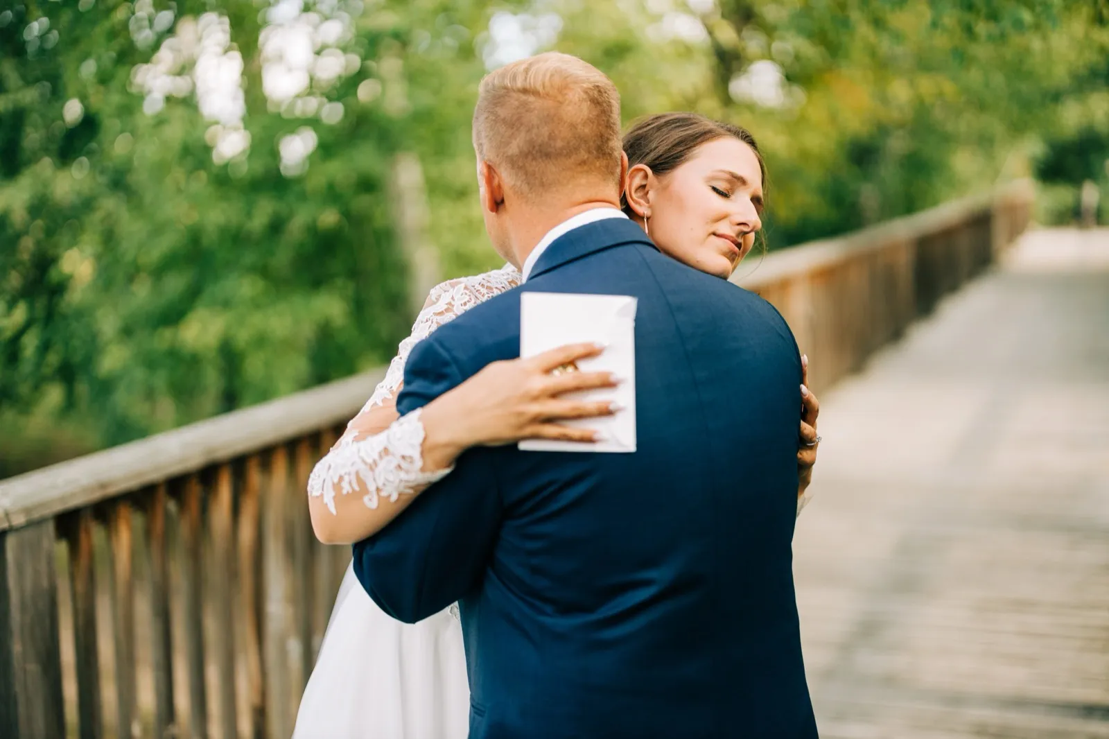 The bride rests her face against the groom's shoulder with eyes closed, holding a folded vow card against his back as they embrace on a wooden footbridge. — Tim Larsen Photography, Brainerd Lakes MN