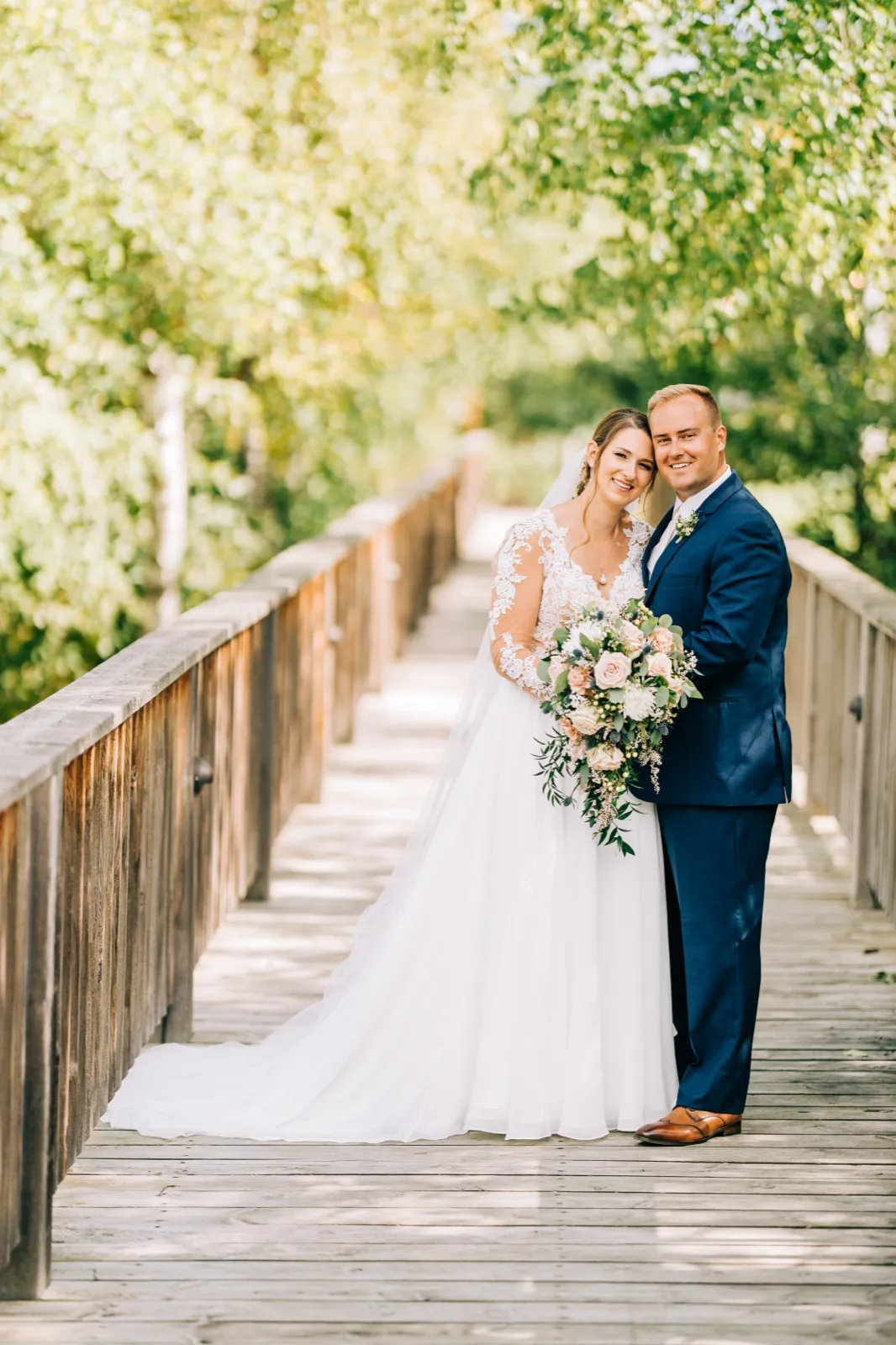 The bride and groom stand together on a wooden footbridge surrounded by green foliage, the bride holding a blush and white bouquet with her train spread behind her. — Tim Larsen Photography, Brainerd Lakes MN