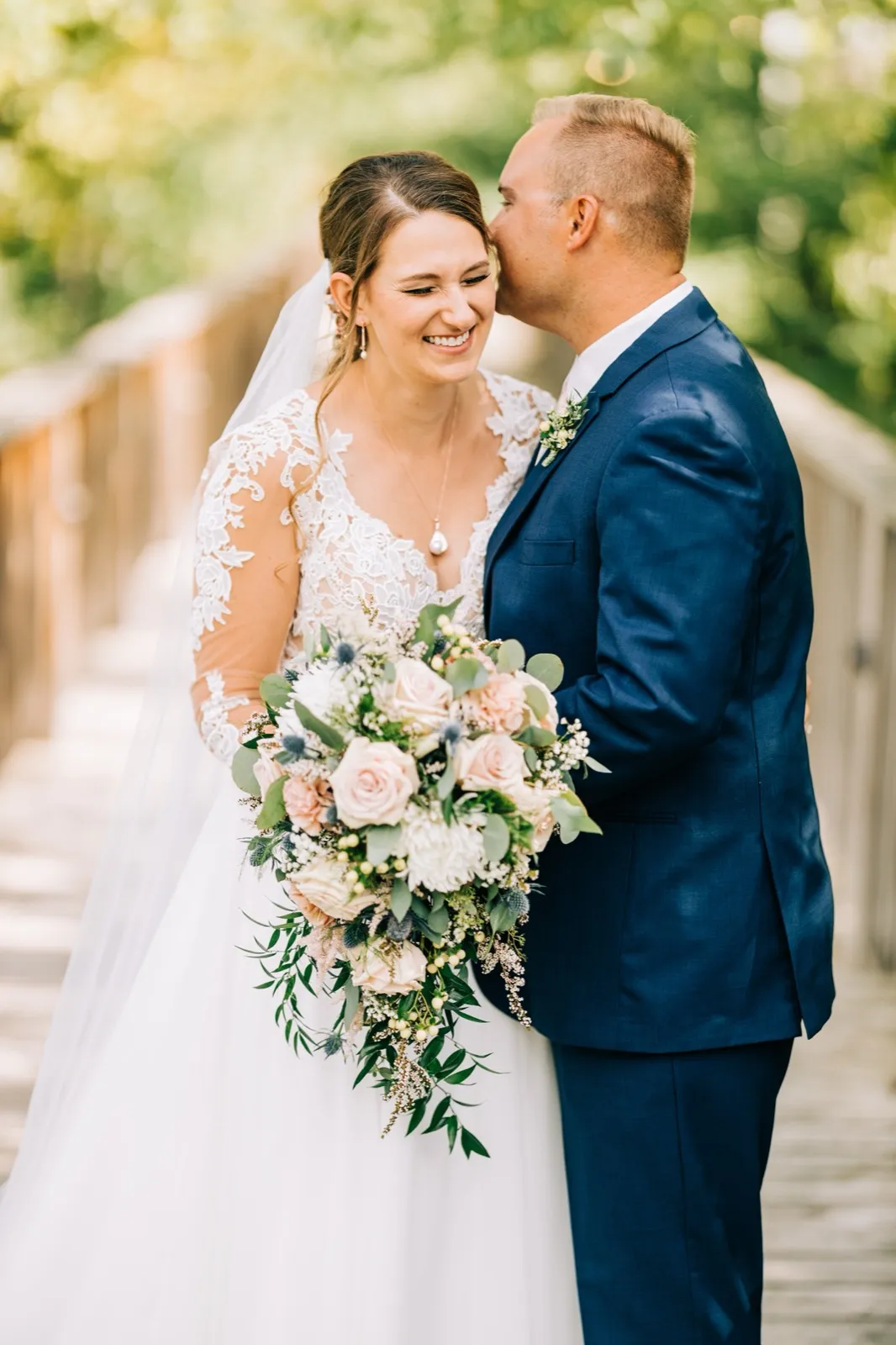 The groom kisses the bride's temple as she laughs, holding a cascading blush and white bouquet on a wooden bridge framed by greenery. — Tim Larsen Photography, Brainerd Lakes MN