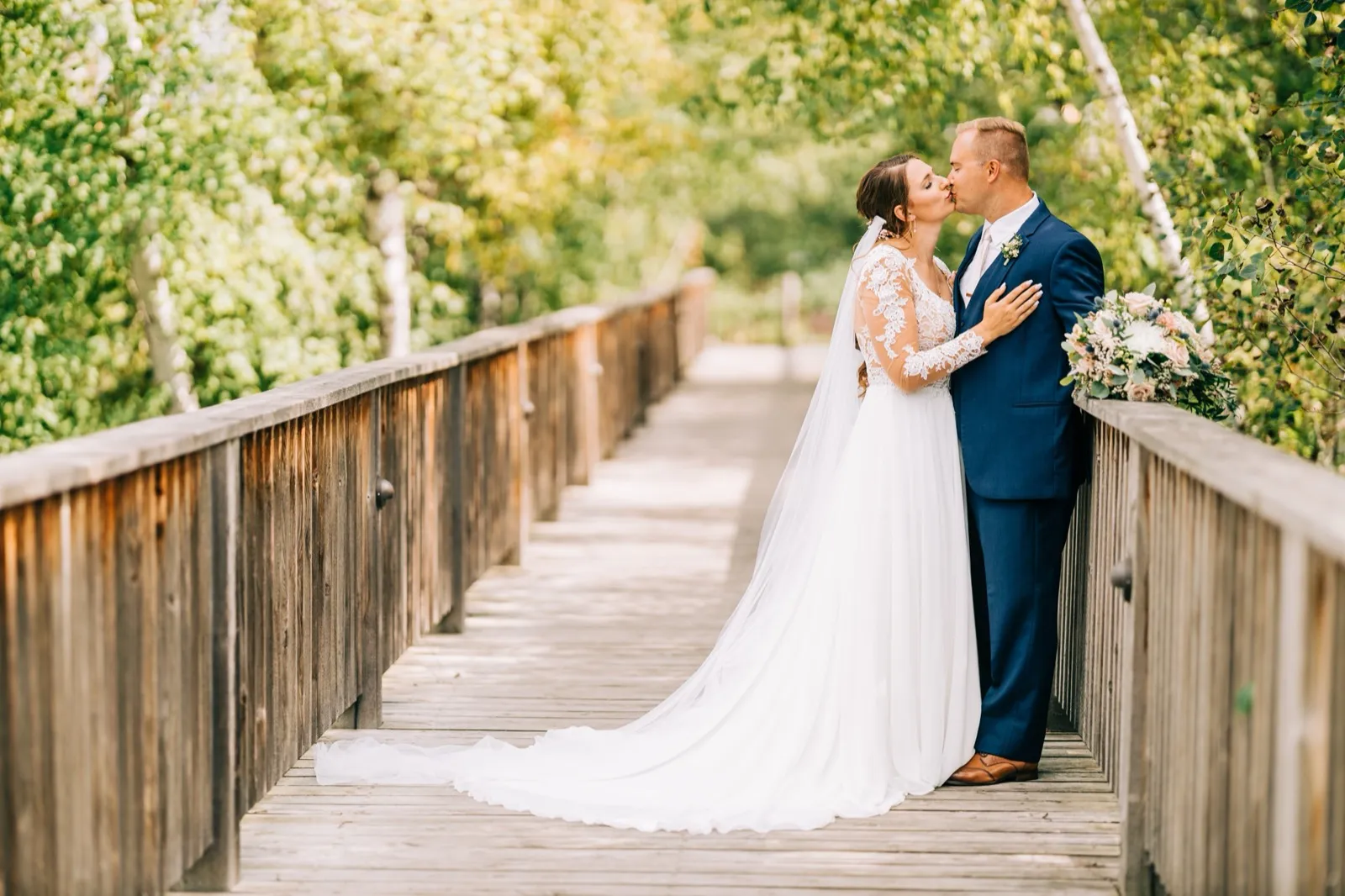 The bride and groom kiss at the center of a wooden footbridge surrounded by birch trees, her veil and dress train spilling across the planks. — Tim Larsen Photography, Brainerd Lakes MN