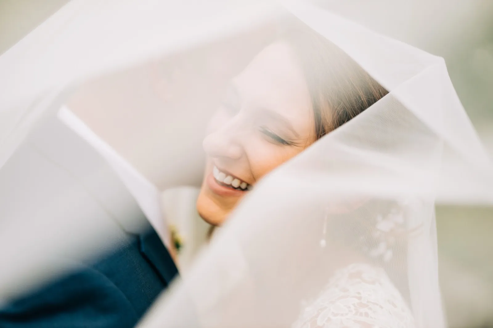 The bride laughs with her eyes closed beneath a translucent veil that drapes over her and the groom during an outdoor portrait. — Tim Larsen Photography, Brainerd Lakes MN