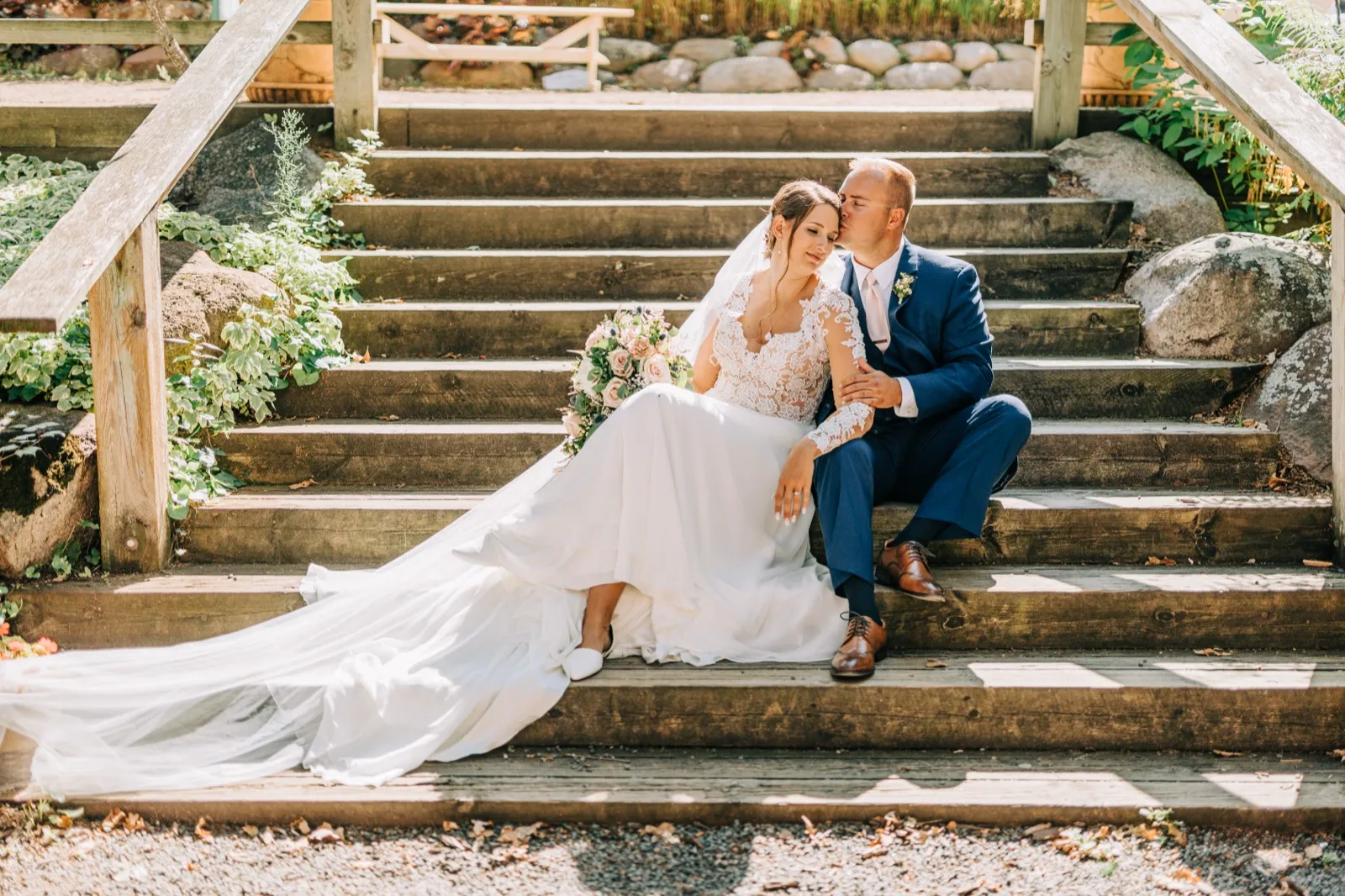 The groom kisses the bride's forehead as they sit together on a wooden stairway, her dress and train spilling down the steps with a bouquet at her side. — Tim Larsen Photography, Brainerd Lakes MN